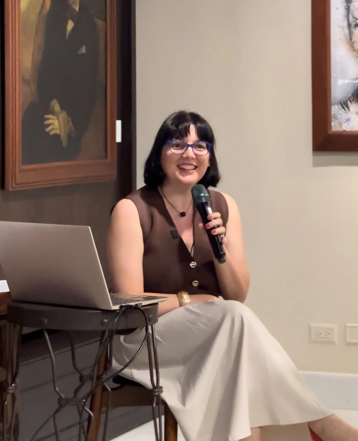 Mujer con gafas negras, camiseta marrón, pantalones blancos, sosteniendo un micrófono y sonriendo, sentada junto a una laptop, en una galería de arte con pinturas en las paredes.