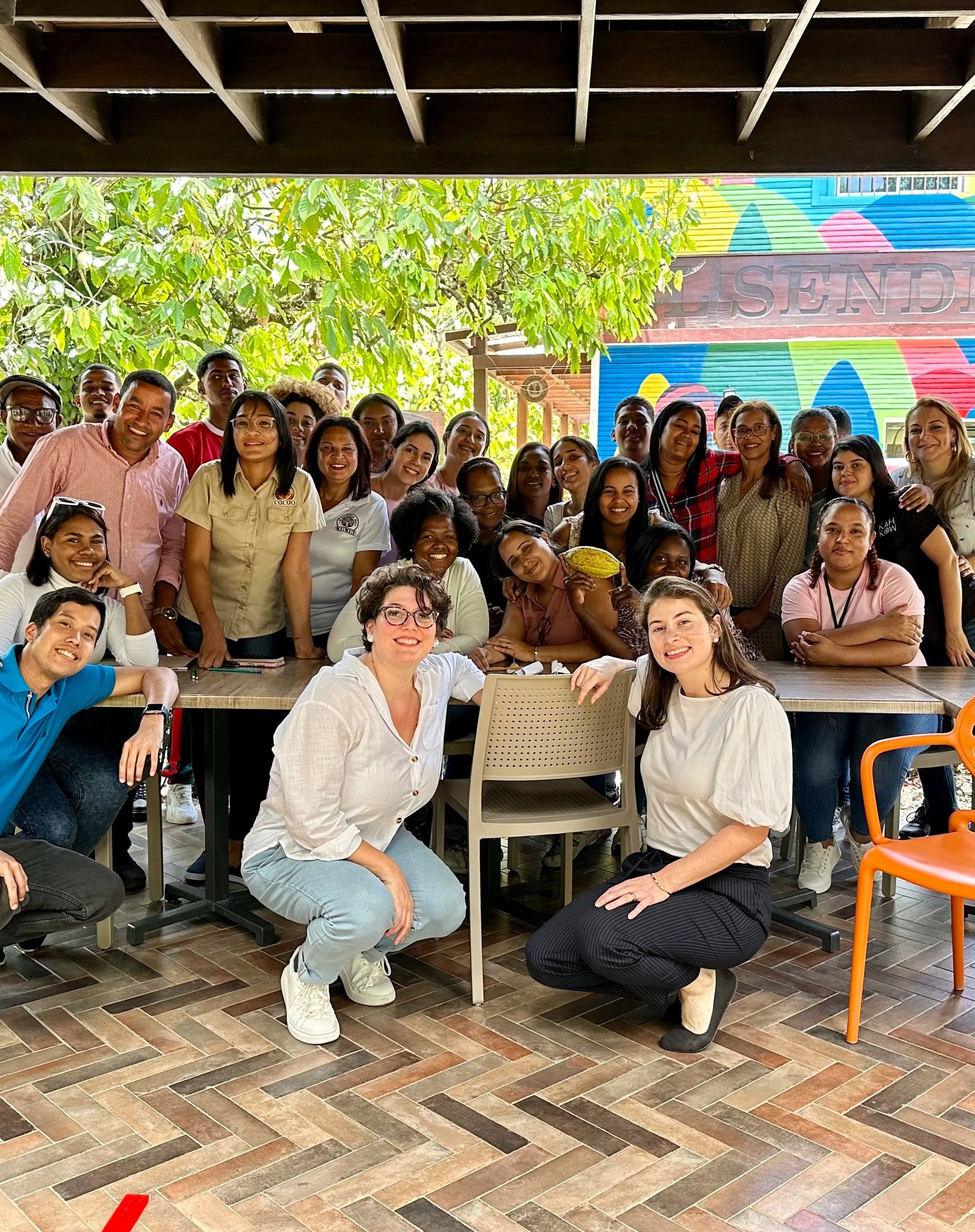 Grupo grande de personas, algunas sonrientes, en un espacio interior con vista a un área exterior con árboles y un muro decorado en colores brillantes.