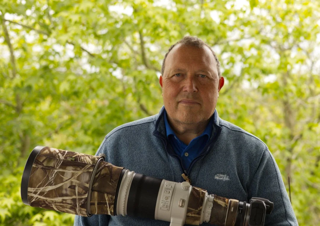 Rob Scotcher holding his camera in a forested area with green leaves.