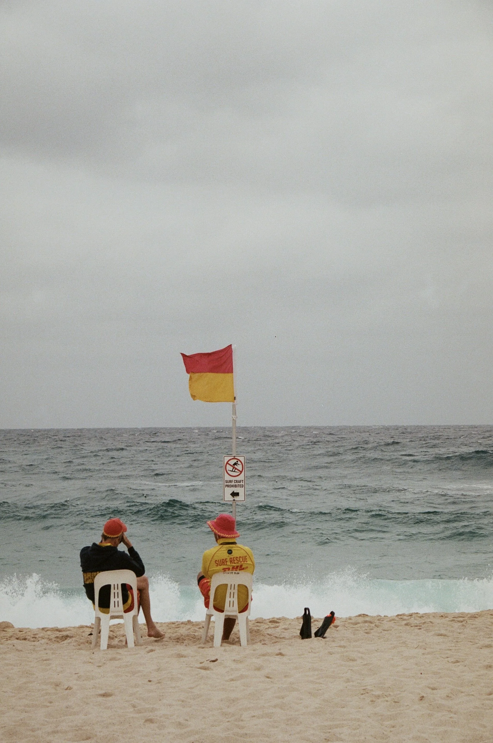 People sitting on chairs on the beach, watching the ocean, with a yellow and red flag and a sign indicating that surf craft is prohibited.