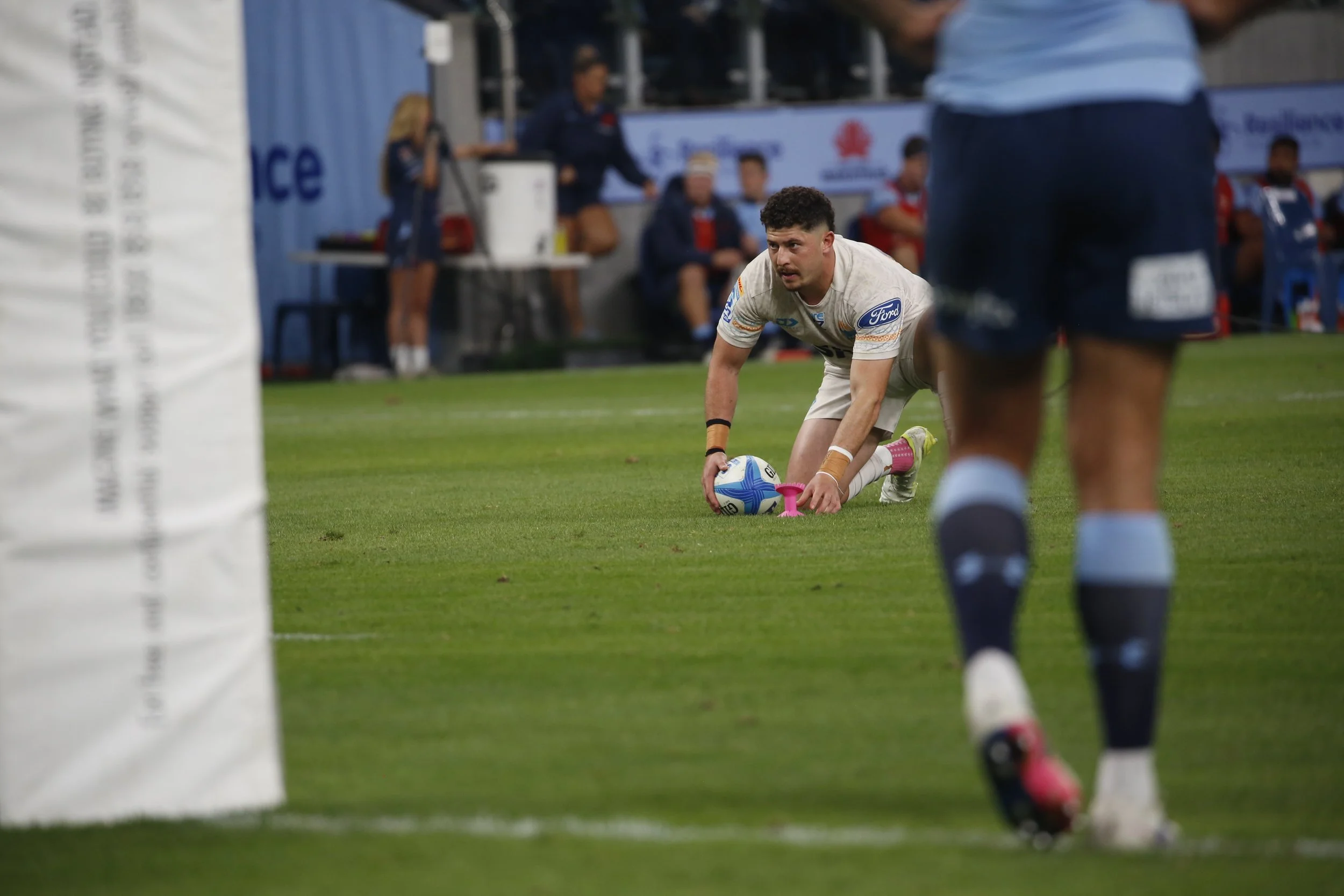 A rugby player with a mustache and curly hair is on his hands and knees on the grass field, holding a rugby ball, during a game. Other players and team staff are in the background, some sitting on the bench and others standing, watching the game.
