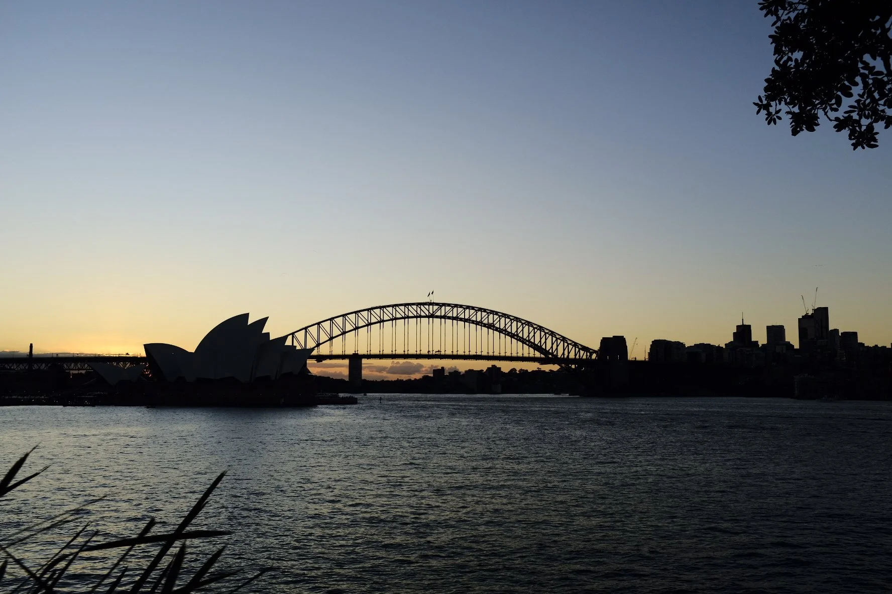Silhouette of the Sydney Opera House and Harbour Bridge at sunset, with city skyline in the background and calm water in foreground.