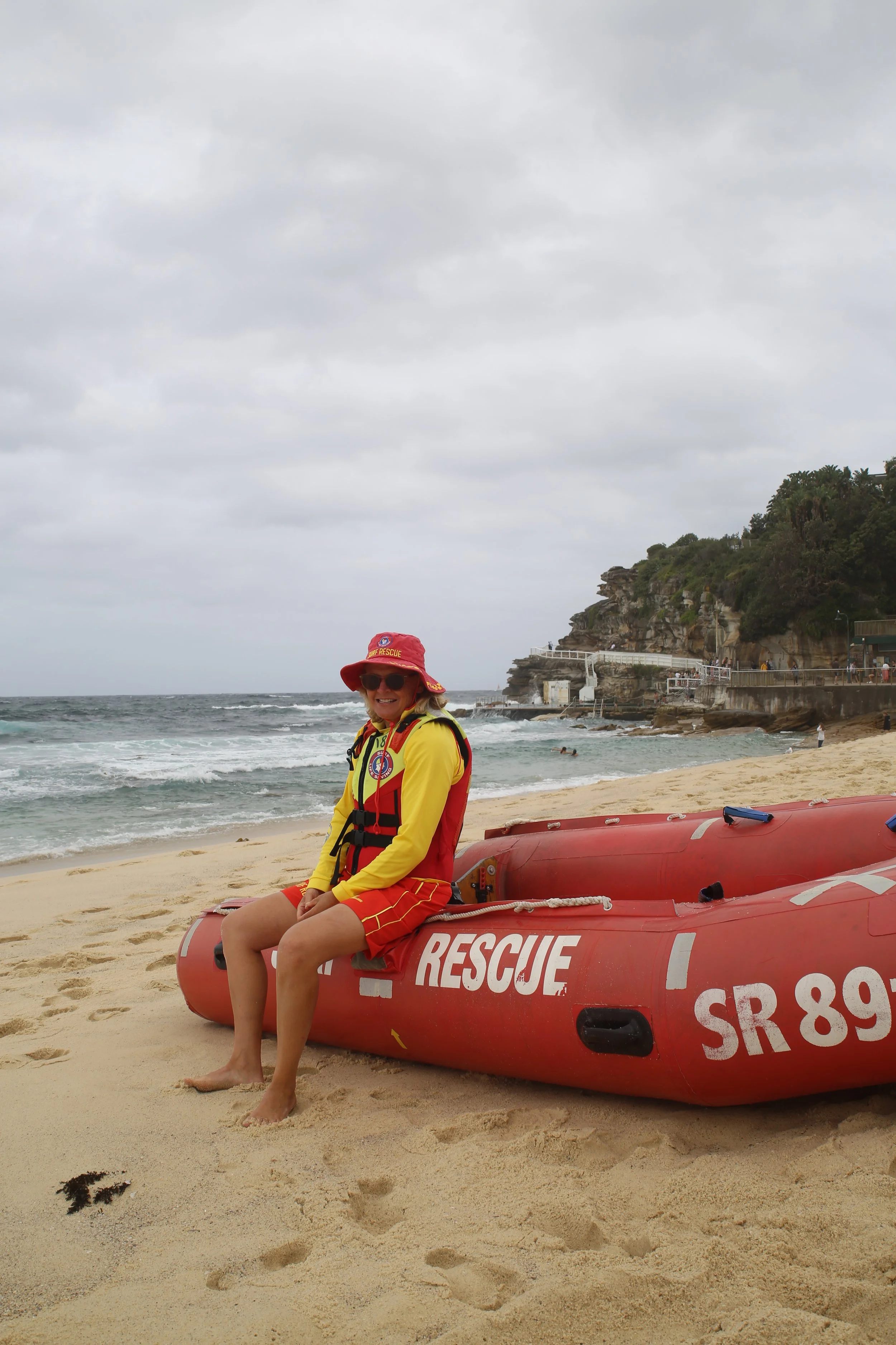 A woman in a yellow rescue uniform with a red rescue hat and sunglasses sitting on a red rescue boat on the sandy beach, with the ocean and rocky cliffs in the background on a cloudy day.