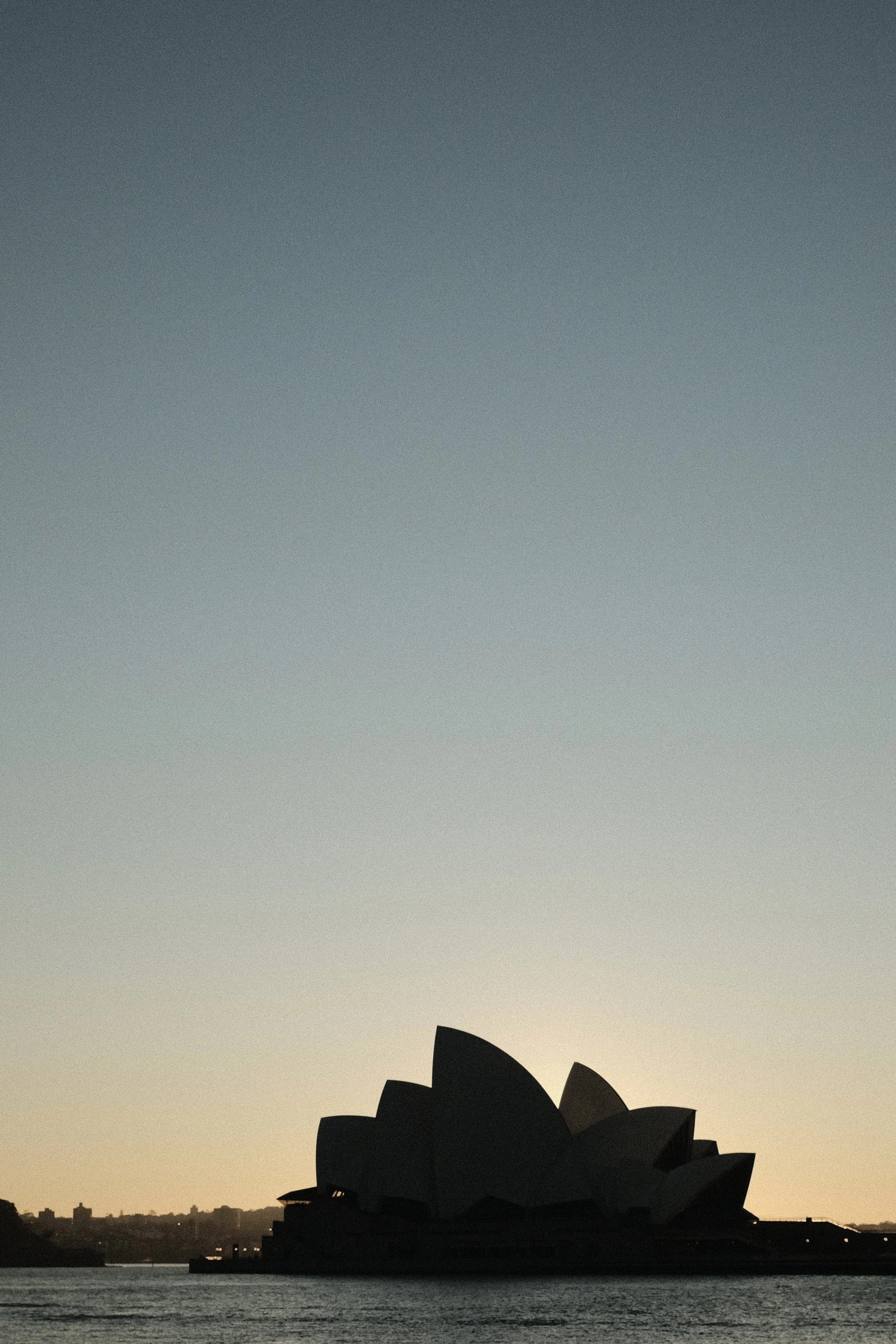 Silhouette of the Sydney Opera House at sunset with a colorful sky, water in the foreground, and city skyline in the background.