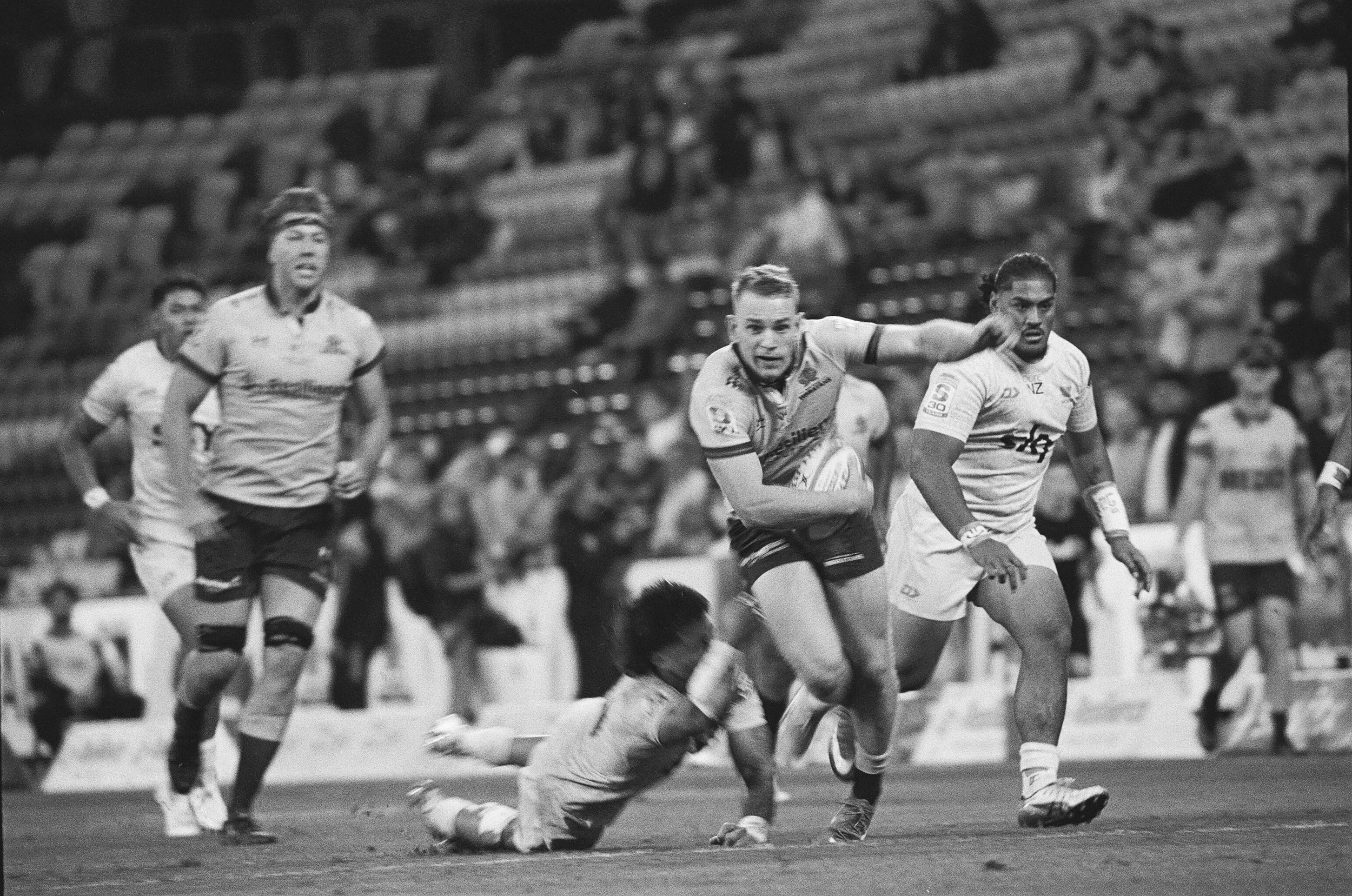 A rugby match in black and white, showing players actively engaged in the game. One player is tackling another who has the ball, while teammates and spectators are visible in the background.