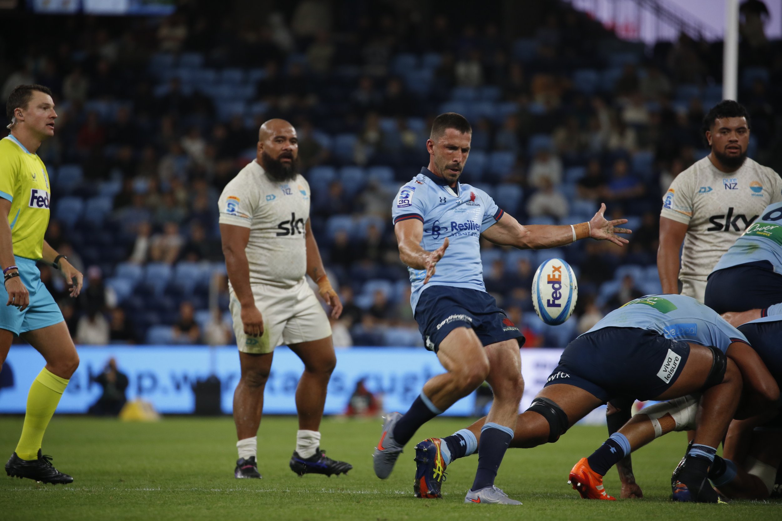 Rugby players in white and light blue jerseys on the field during a game, with crowd in the background.