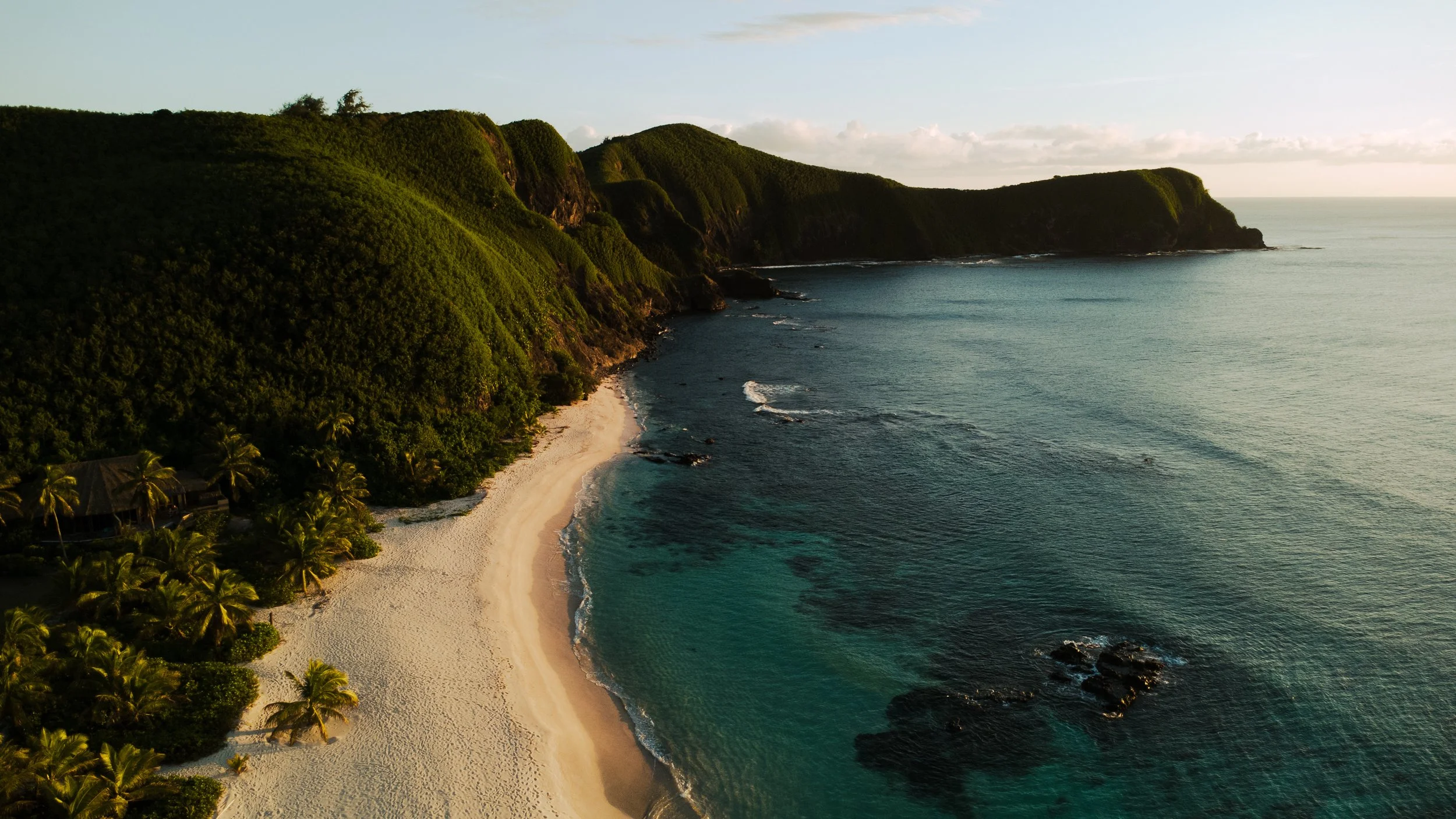 Aerial view of a tropical beach with white sand, lush green cliffs, and ocean waves, illuminated by sunlight.