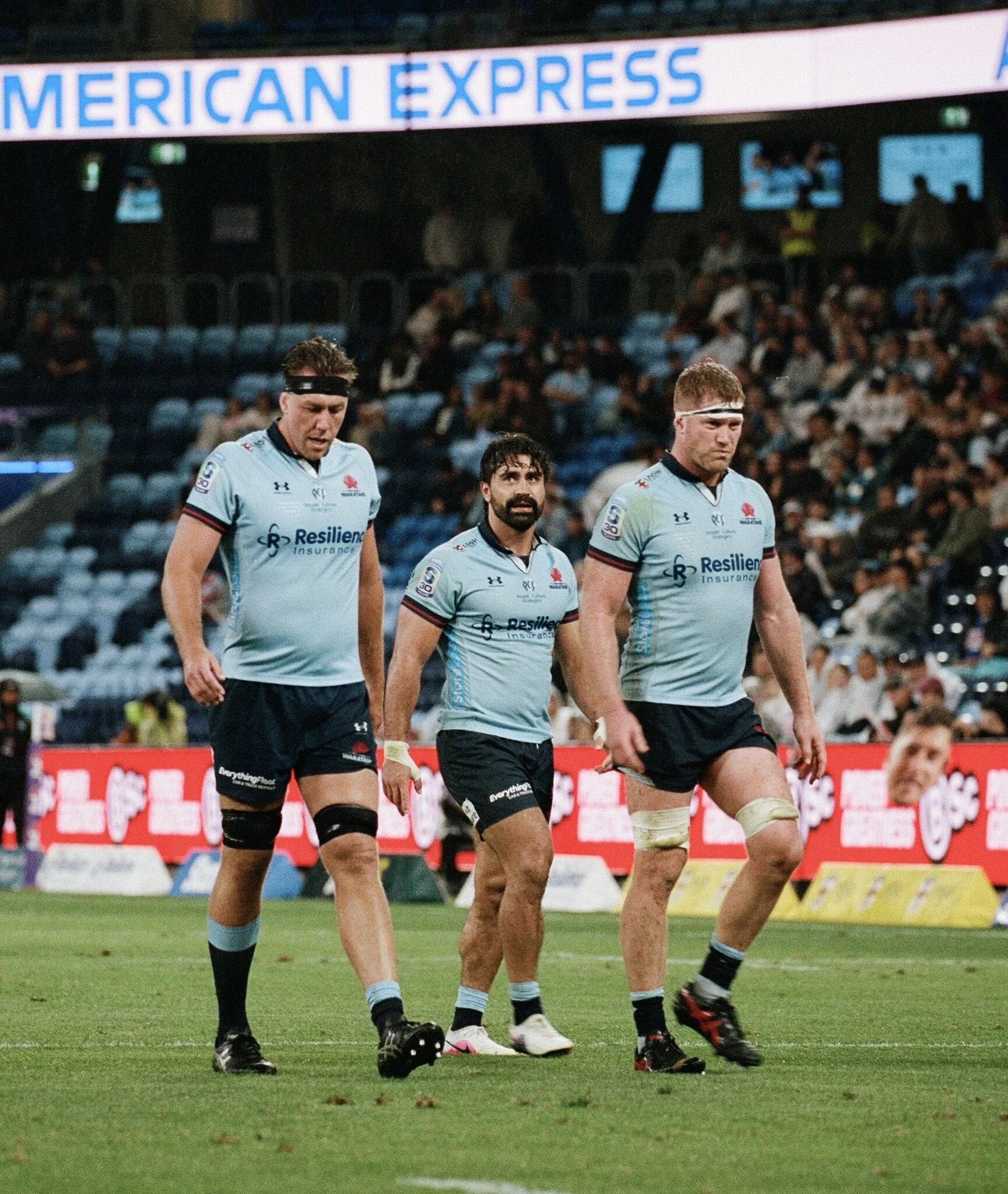 Three rugby players walking on a rugby field during a match, wearing light blue jerseys and black shorts, with a crowd in the background and a large sign that reads 'AMERICAN EXPRESS'.
