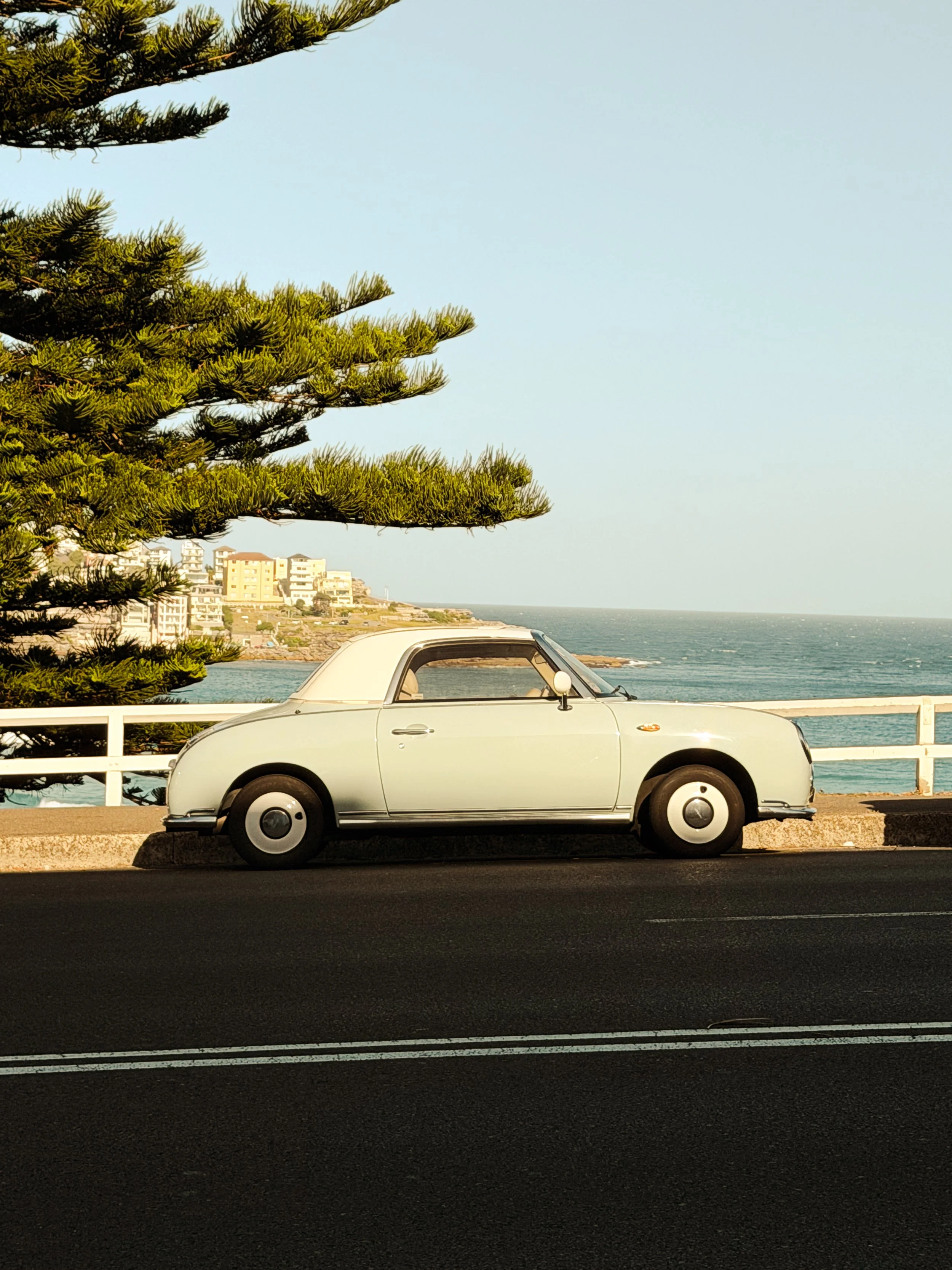 A vintage white convertible car parked on a street near the ocean, with a tree, white railing, and a coastal town in the background.