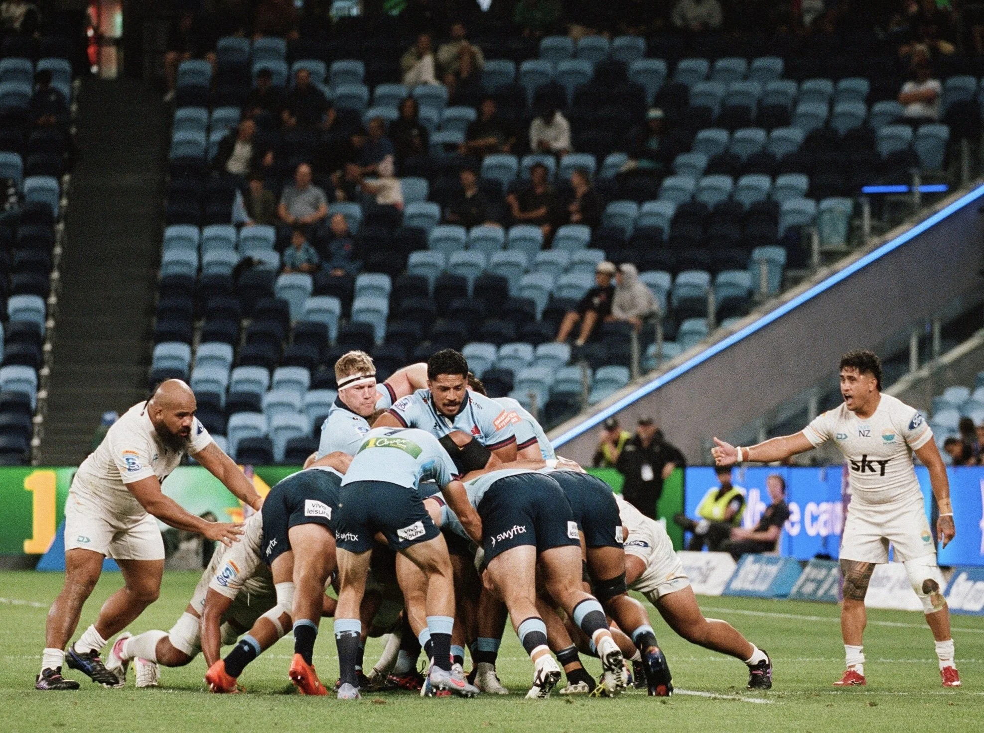 Rugby players engaged in a scrum during a match on a stadium field, with spectators watching in the background.