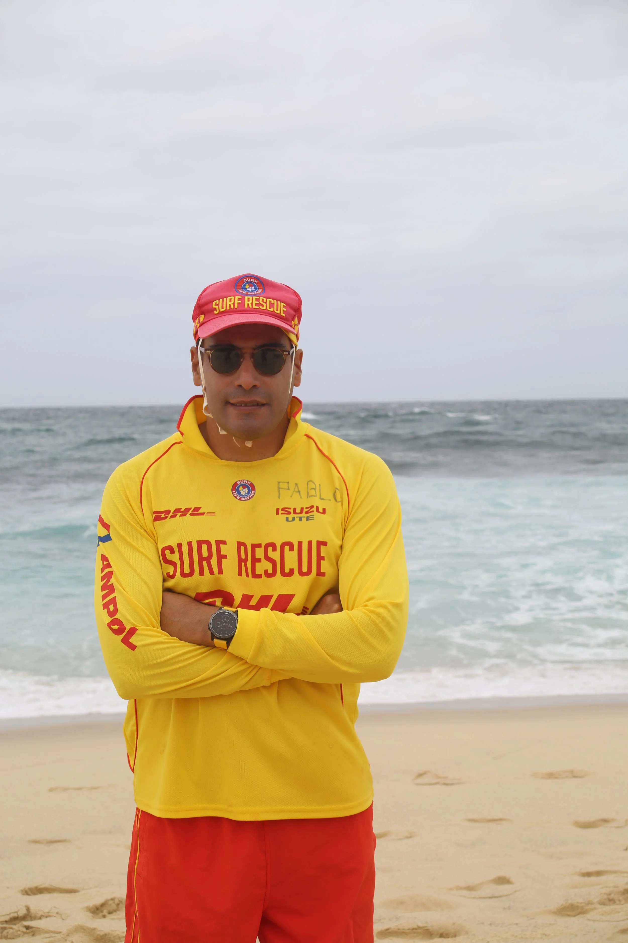 A man wearing a yellow surf rescue shirt and red shorts stands on a beach with the ocean and cloudy sky in the background, crossing his arms and looking at the camera.