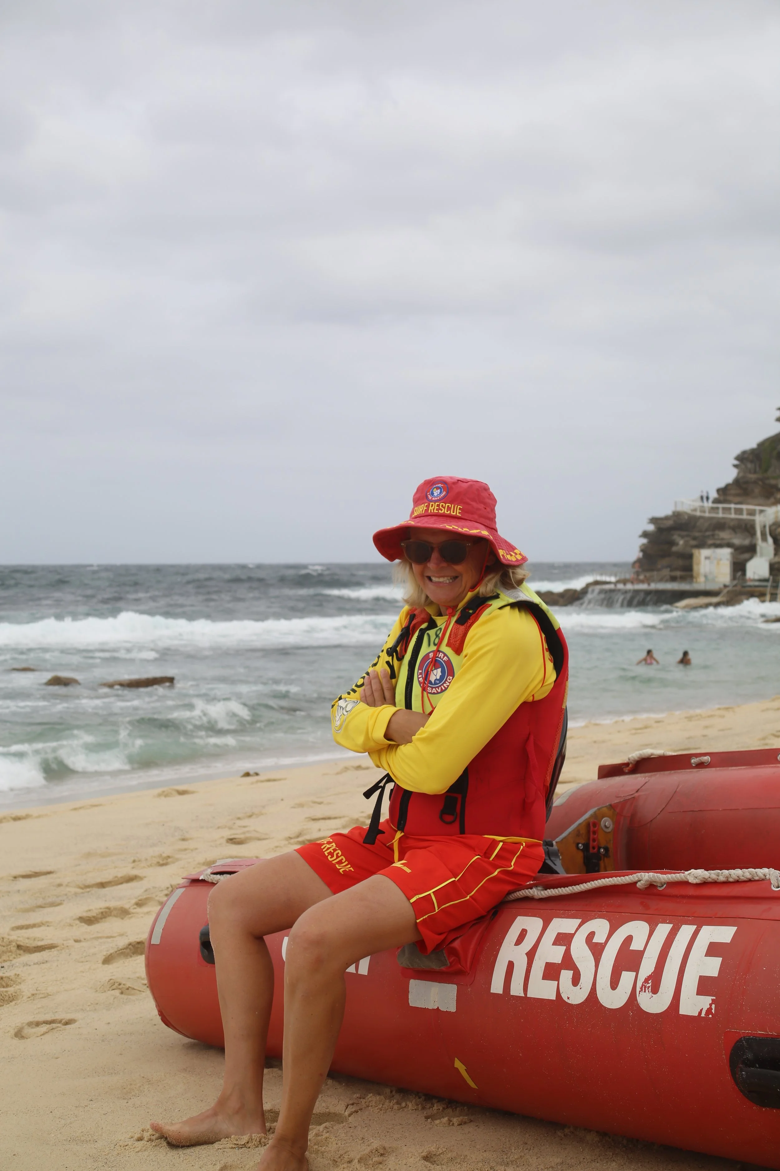 A woman wearing a rescue uniform and a red rescue hat sitting on a red rescue boat on a sandy beach, with the ocean and a rocky cliff in the background.