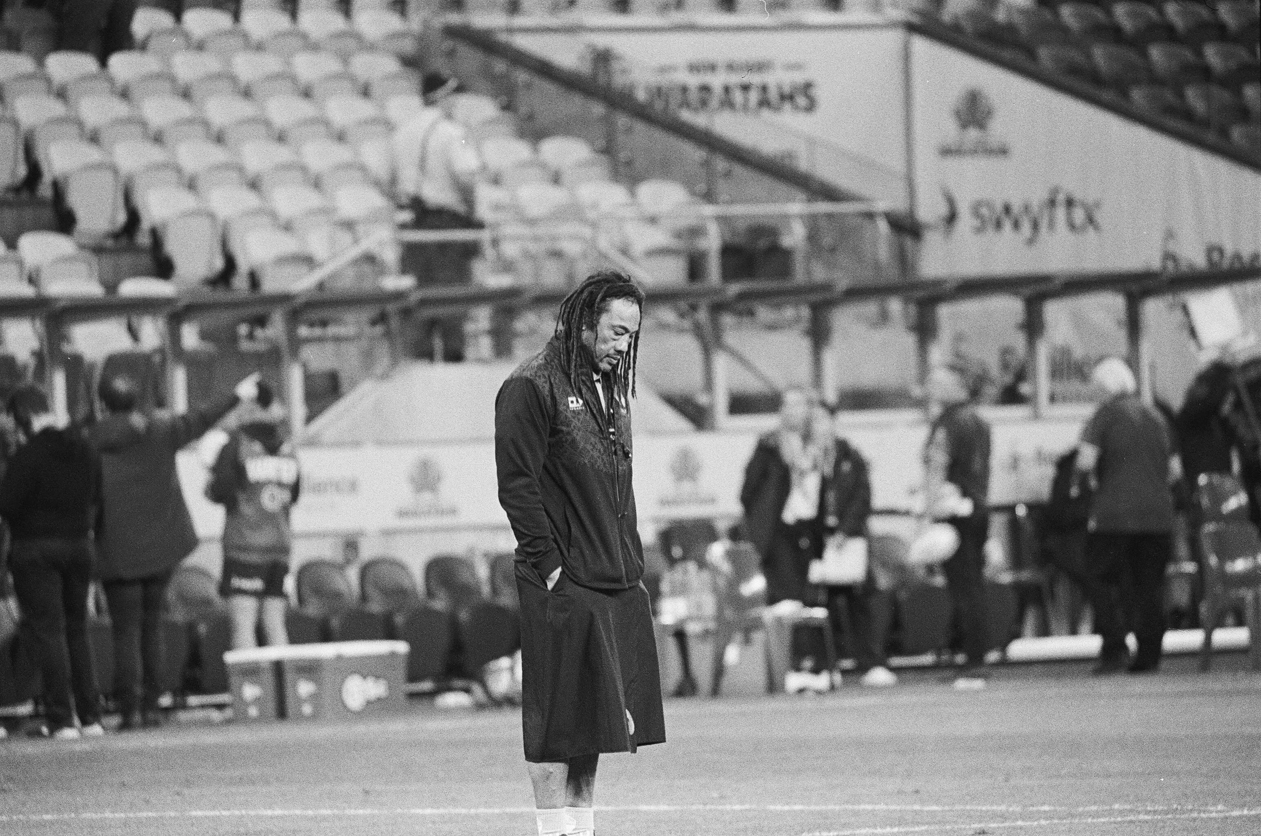 A man with long dreadlocks and a sports jacket standing with his head down on a sports field, with empty stadium seats in the background.