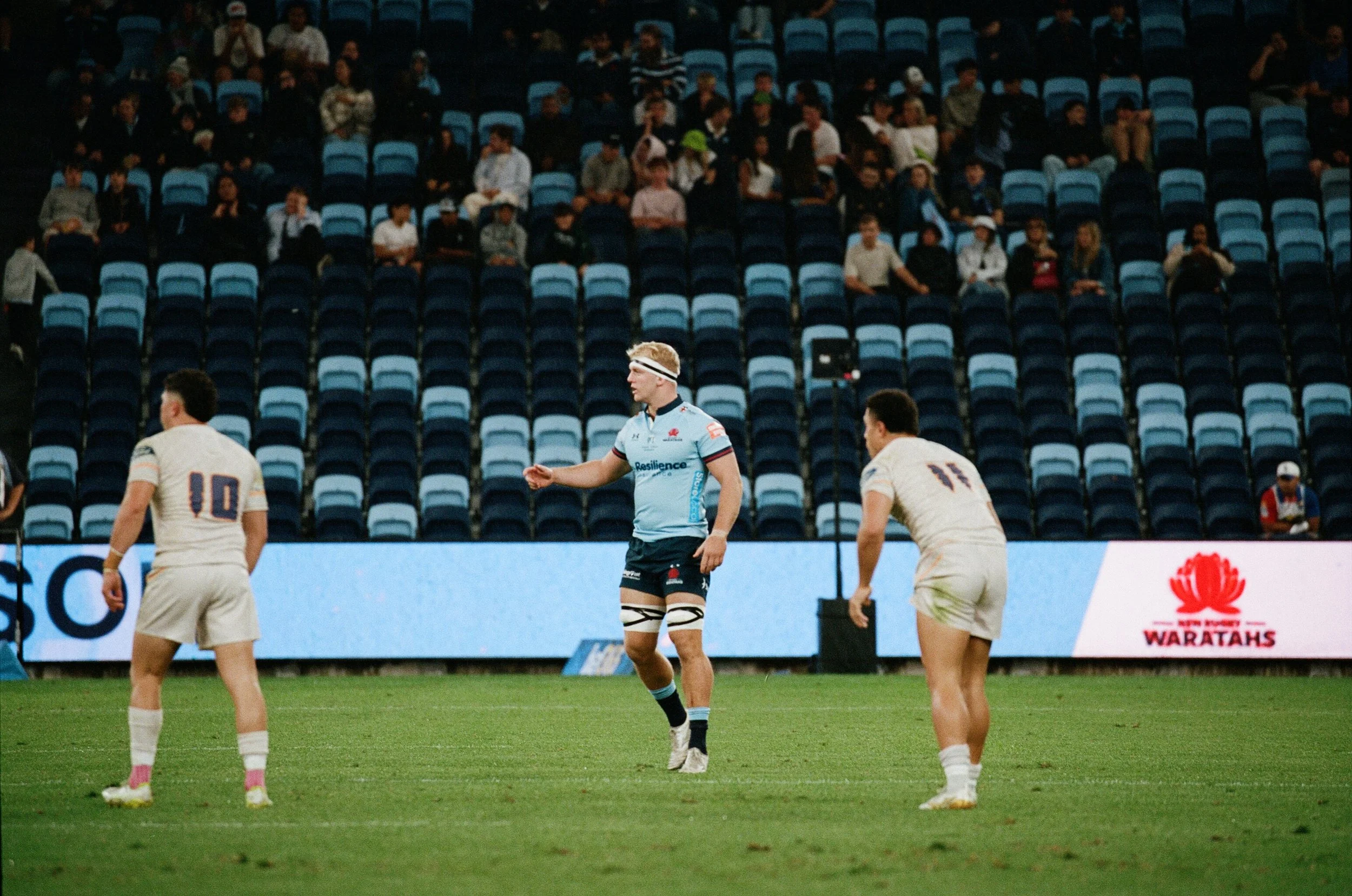 A rugby game in progress with players on the field and spectators seated in the stands.