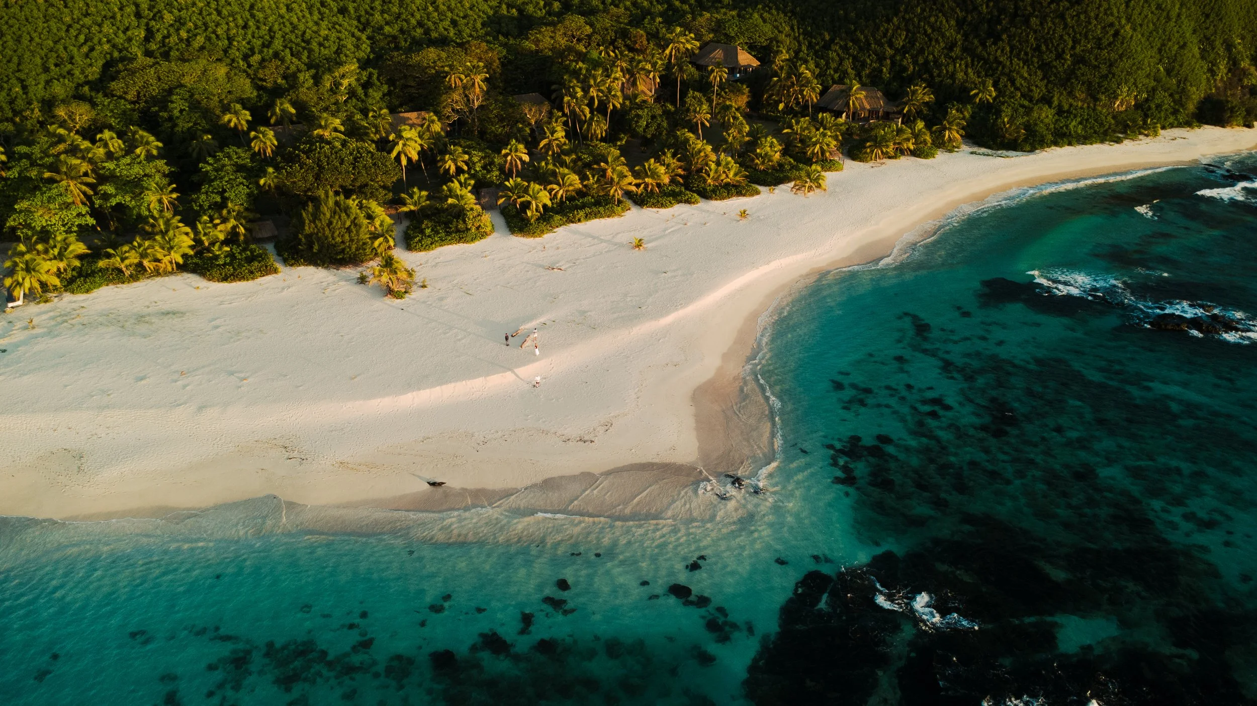 Aerial view of a tropical beach with white sand, turquoise water, and lush green trees with thatched-roof huts, and a few people walking along the shoreline.