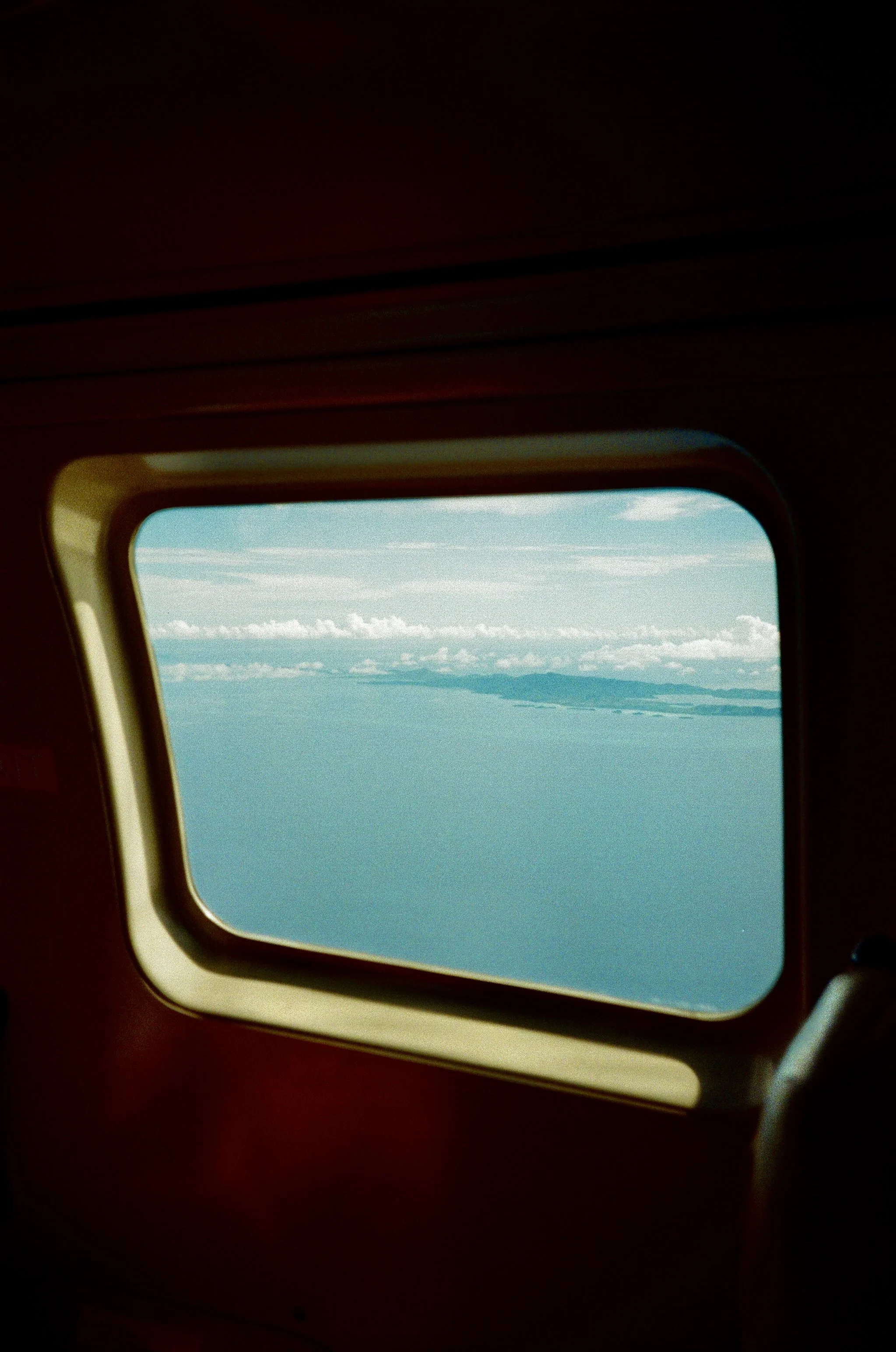 An aircraft window with a view of the sky, clouds, and distant mountains.