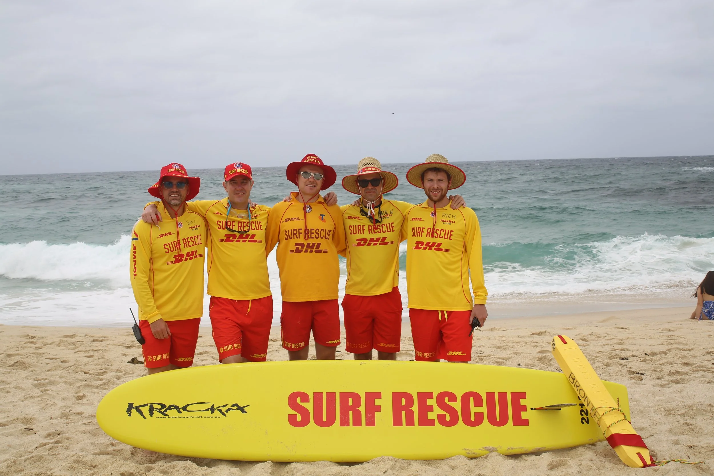 Group of five surf rescue team members standing on the beach in yellow and red uniforms with surf rescue hats, with surfboards and rescue equipment on the sand, ocean waves in the background.