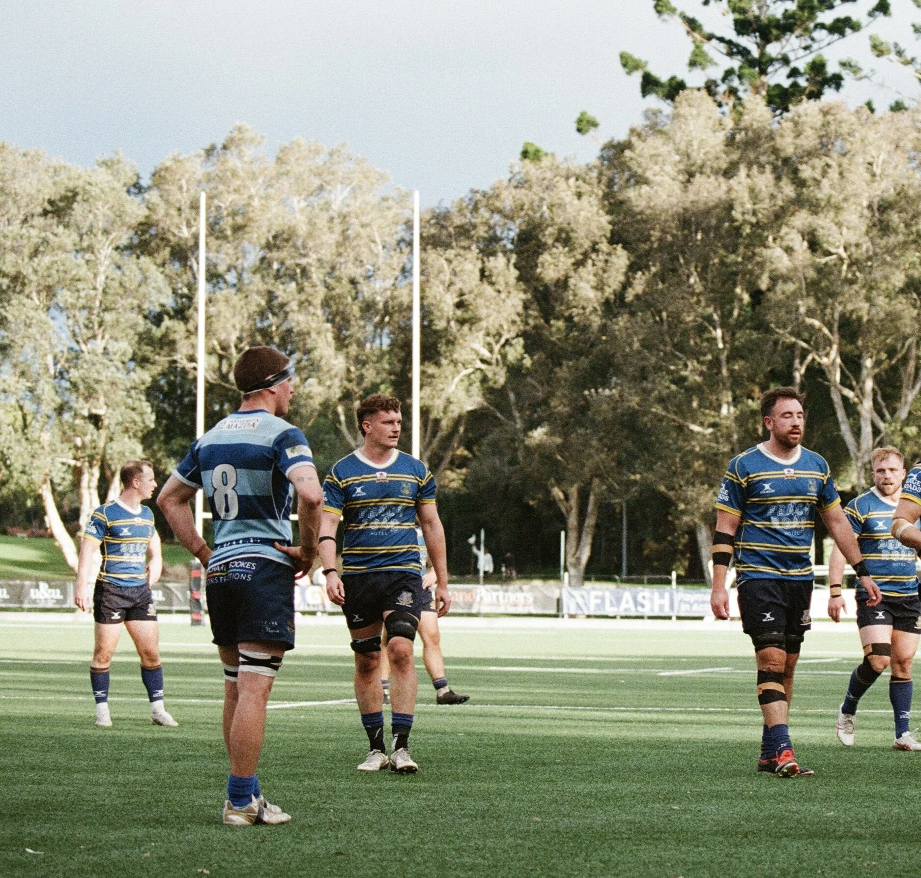 A group of rugby players on the field during a game, wearing blue and yellow uniforms, with trees and goalposts in the background.