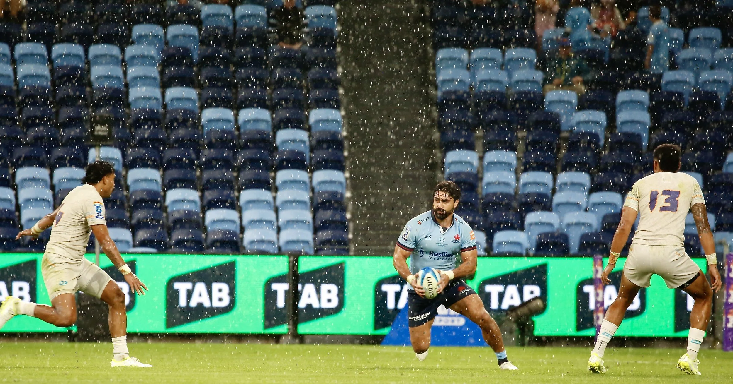 A rugby match taking place in a stadium, rain falling heavily, with players in action and empty blue seats in the background.
