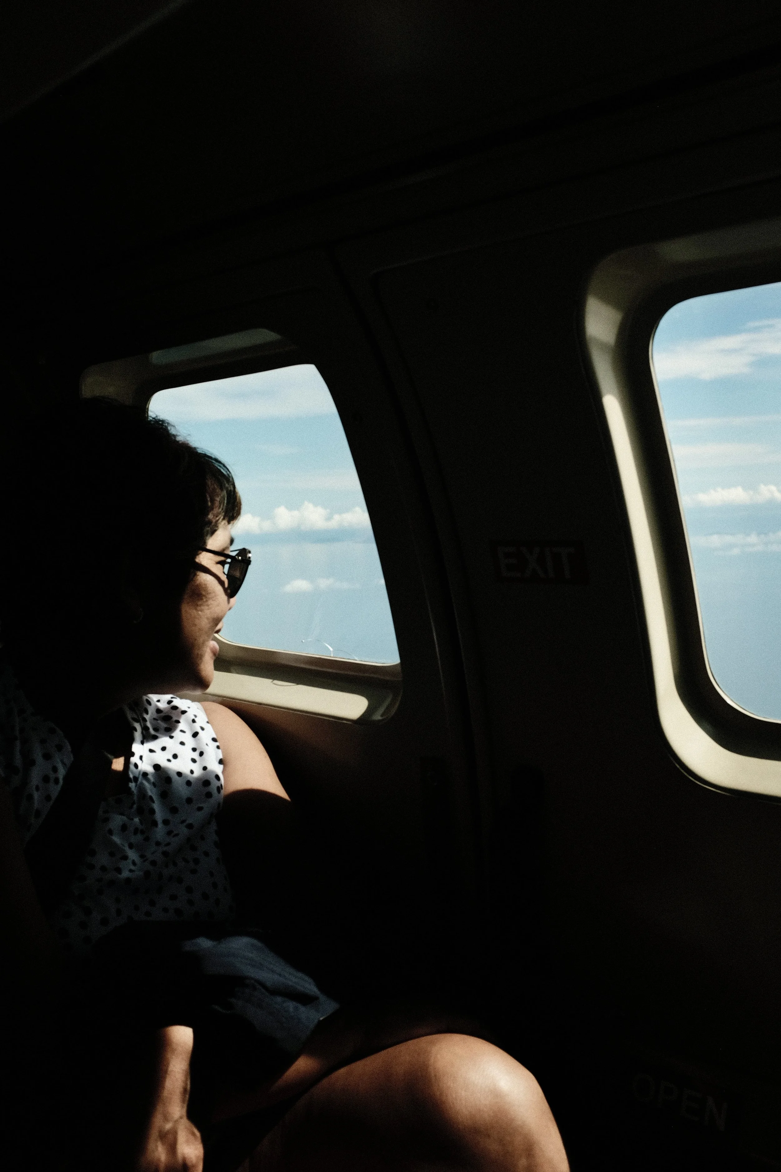 A woman wearing glasses and a polka dot shirt is sitting in a vehicle, looking out of the window at the sky with clouds.