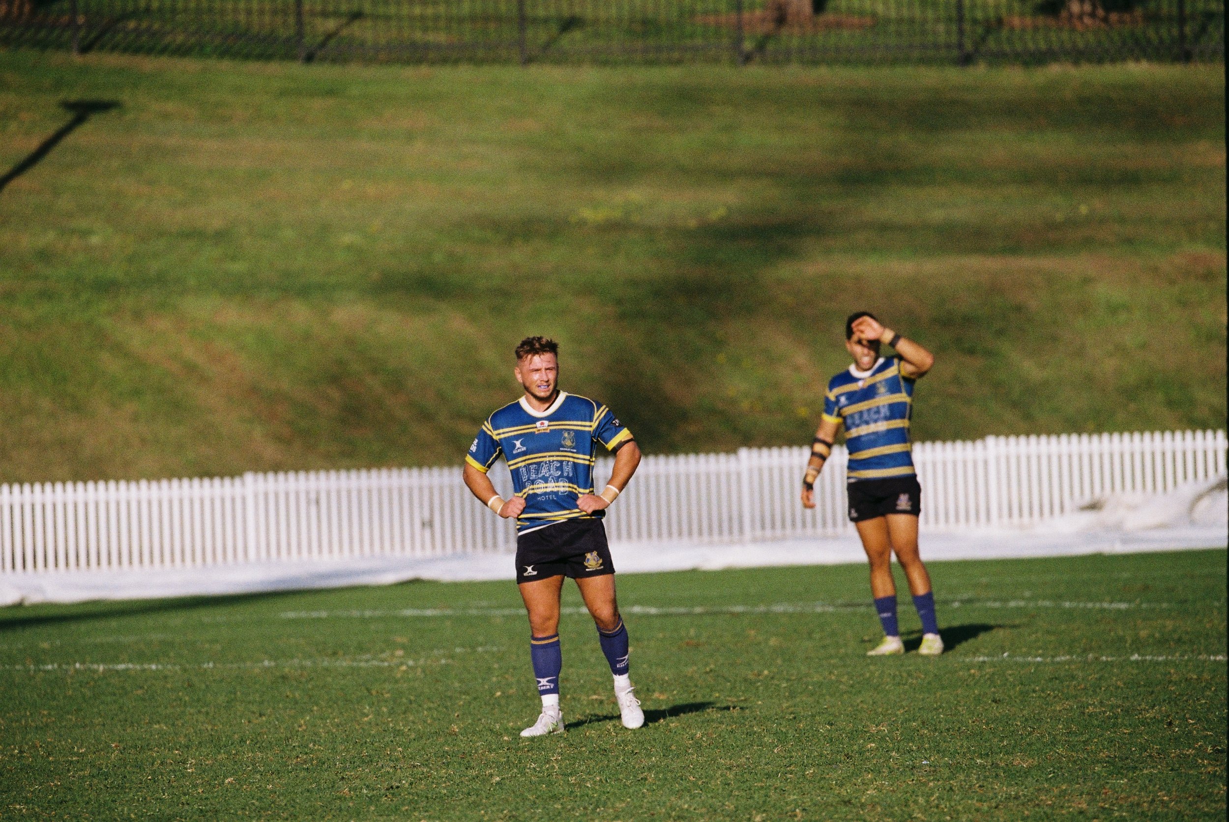 Two rugby players on a field wearing blue and yellow uniforms, one standing with hands on hips and the other with hand on face, behind a white picket fence.