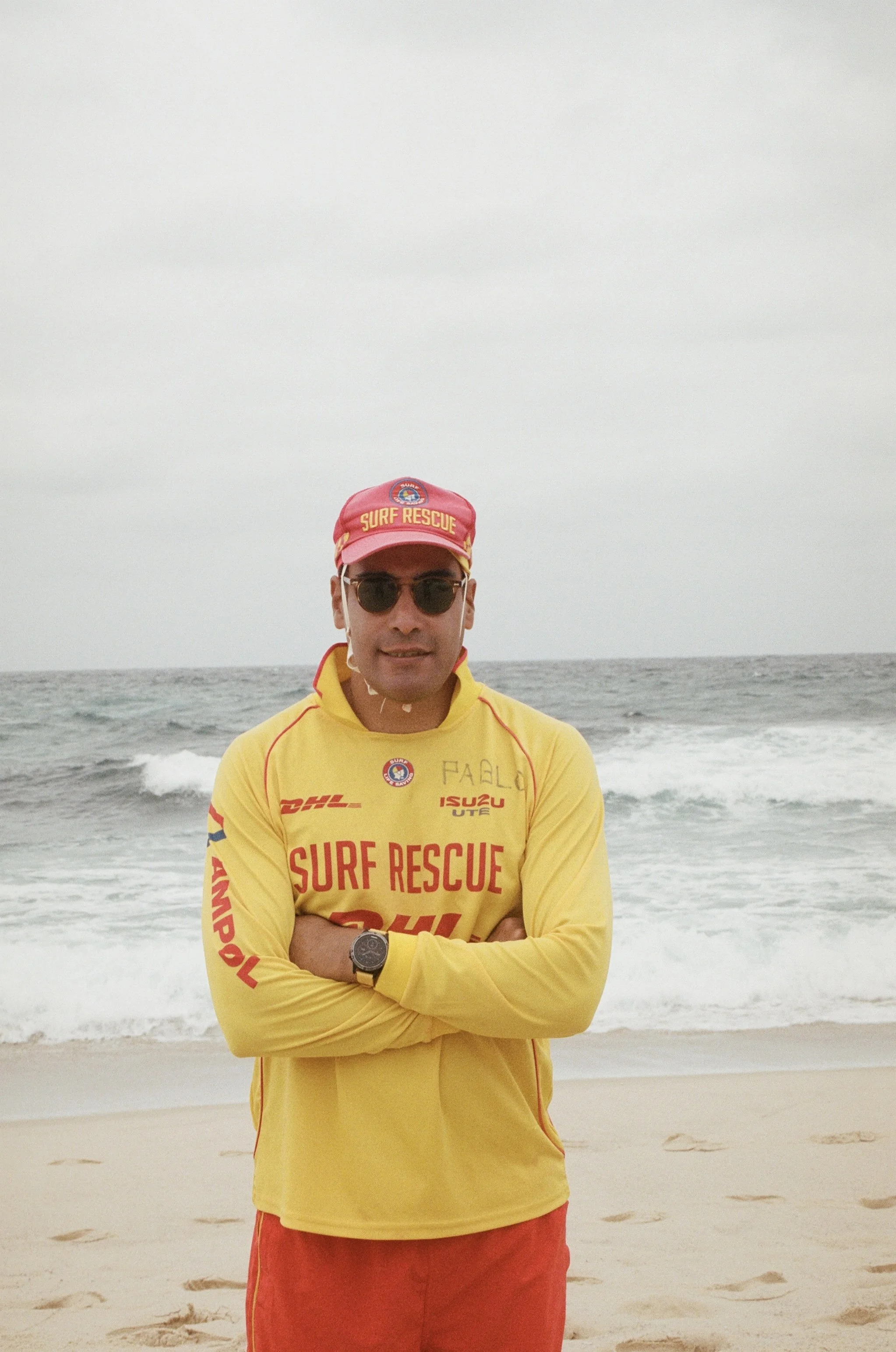 A man wearing sunglasses, a red cap with yellow text reading 'Surf Rescue,' a yellow long-sleeve shirt with red text 'Surf Rescue,' and red shorts, standing on a sandy beach with the ocean and waves in the background.