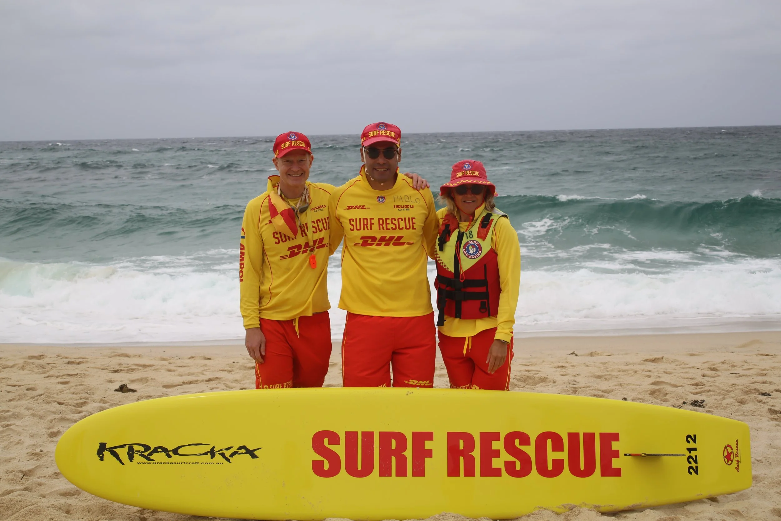 Three surf rescue team members standing on the beach behind a yellow surf rescue board, wearing yellow uniforms with red accents and red caps that say 'Surf Rescue'. They are smiling, and the ocean is visible in the background with overcast skies.