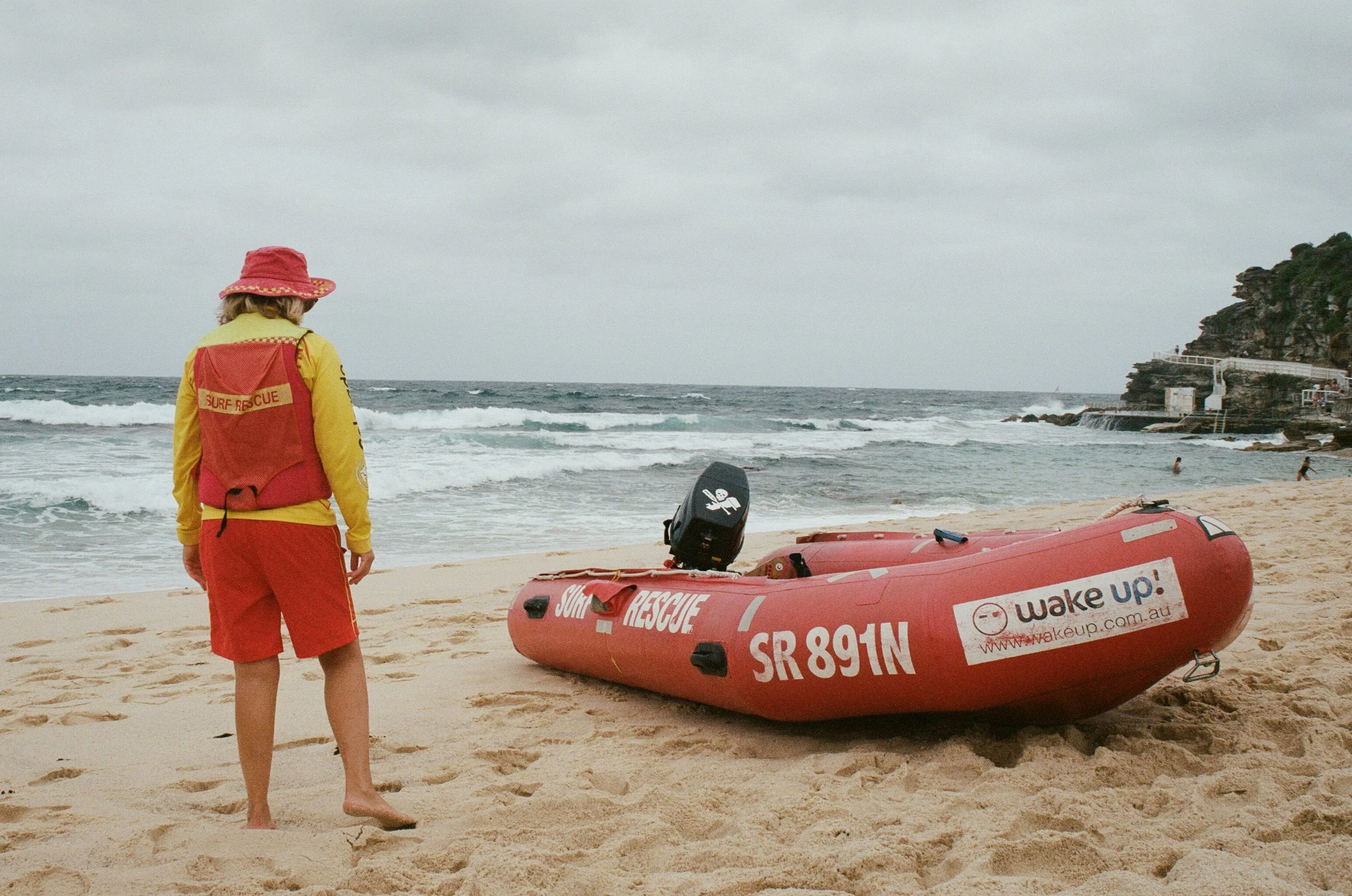 A person wearing a red hat and yellow rescue uniform standing on a sandy beach facing the ocean. There is a red rescue boat and an outboard motor nearby. The sky is overcast, and some people are in the water near the rocky cliffs in the background.