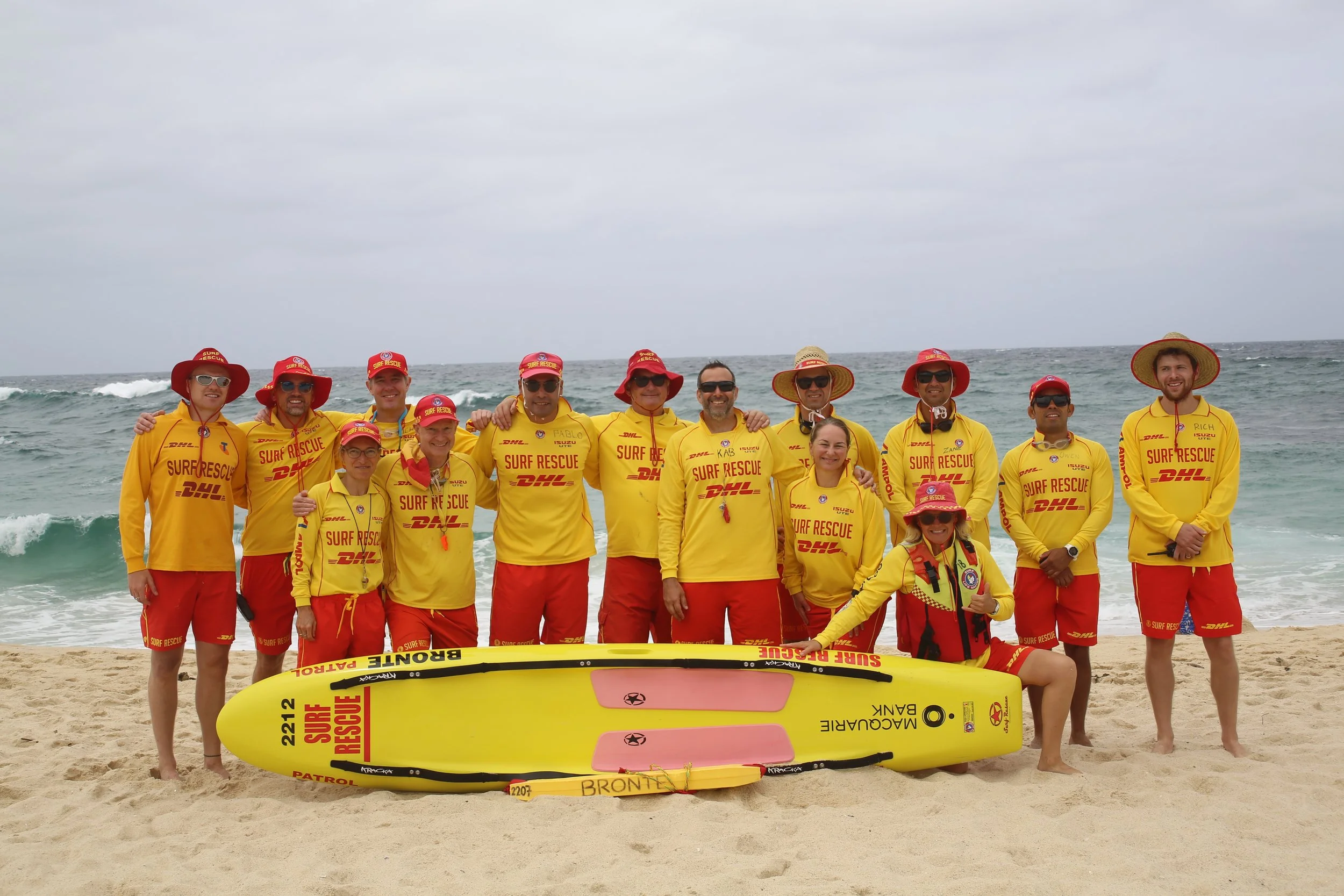 Group of surf rescue team members standing on the beach, wearing yellow uniforms and red hats, with surf rescue equipment and surfboards in front of them, ocean waves in the background.