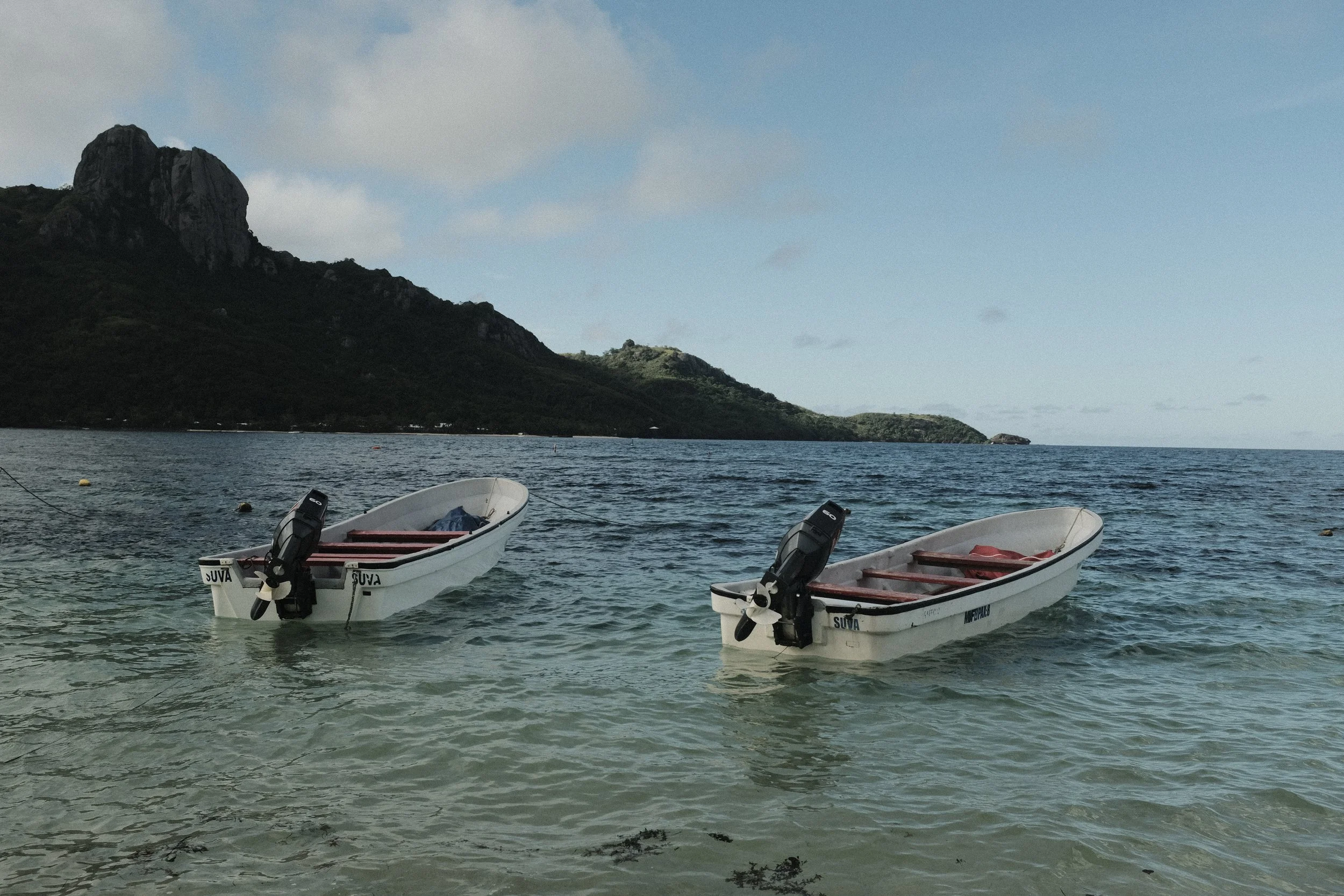Two small white boats with outboard motors floating on the clear water near a rocky island or hillside under a partly cloudy sky.