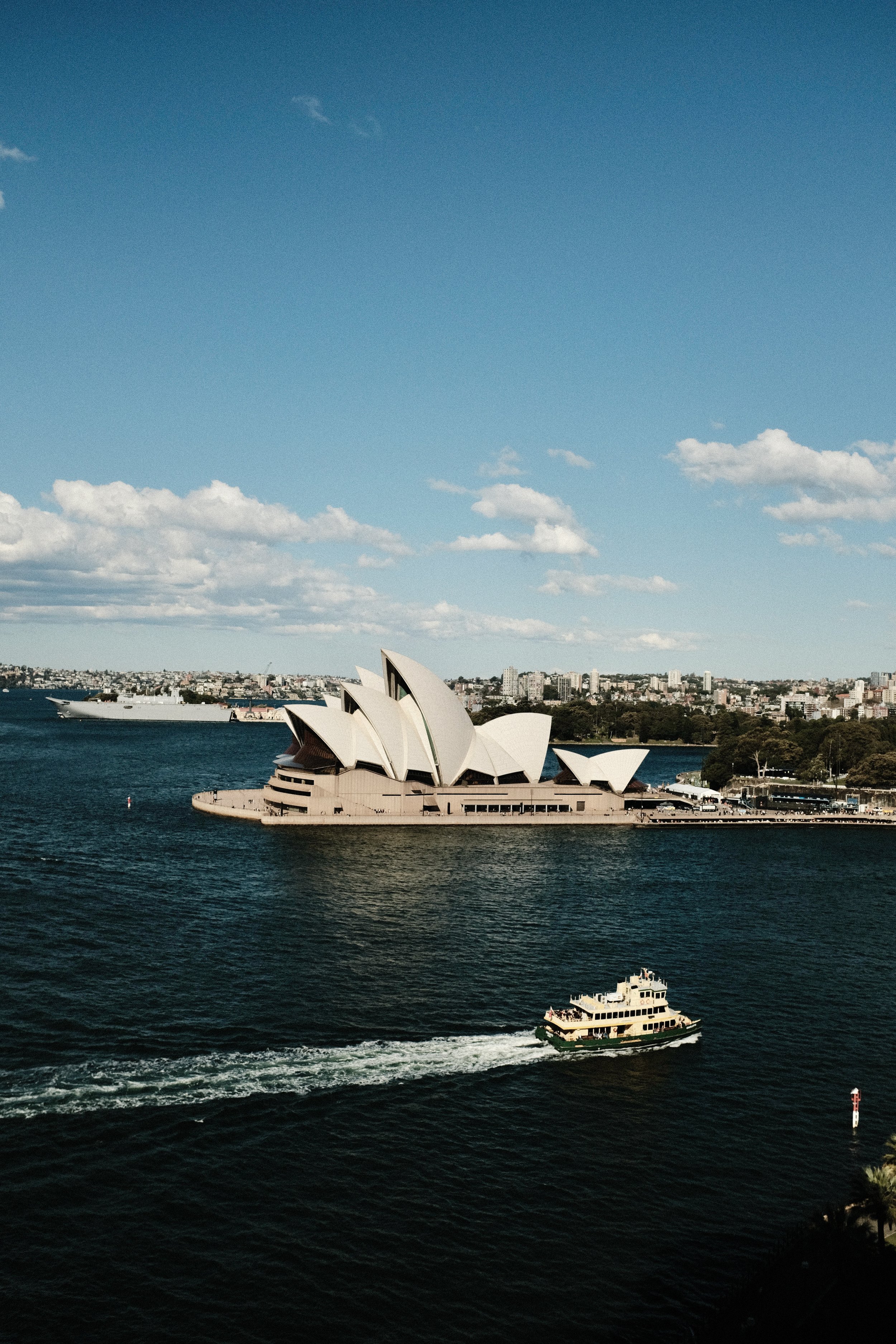 Sydney Opera House with boats on water and city skyline in background under blue sky with scattered clouds.