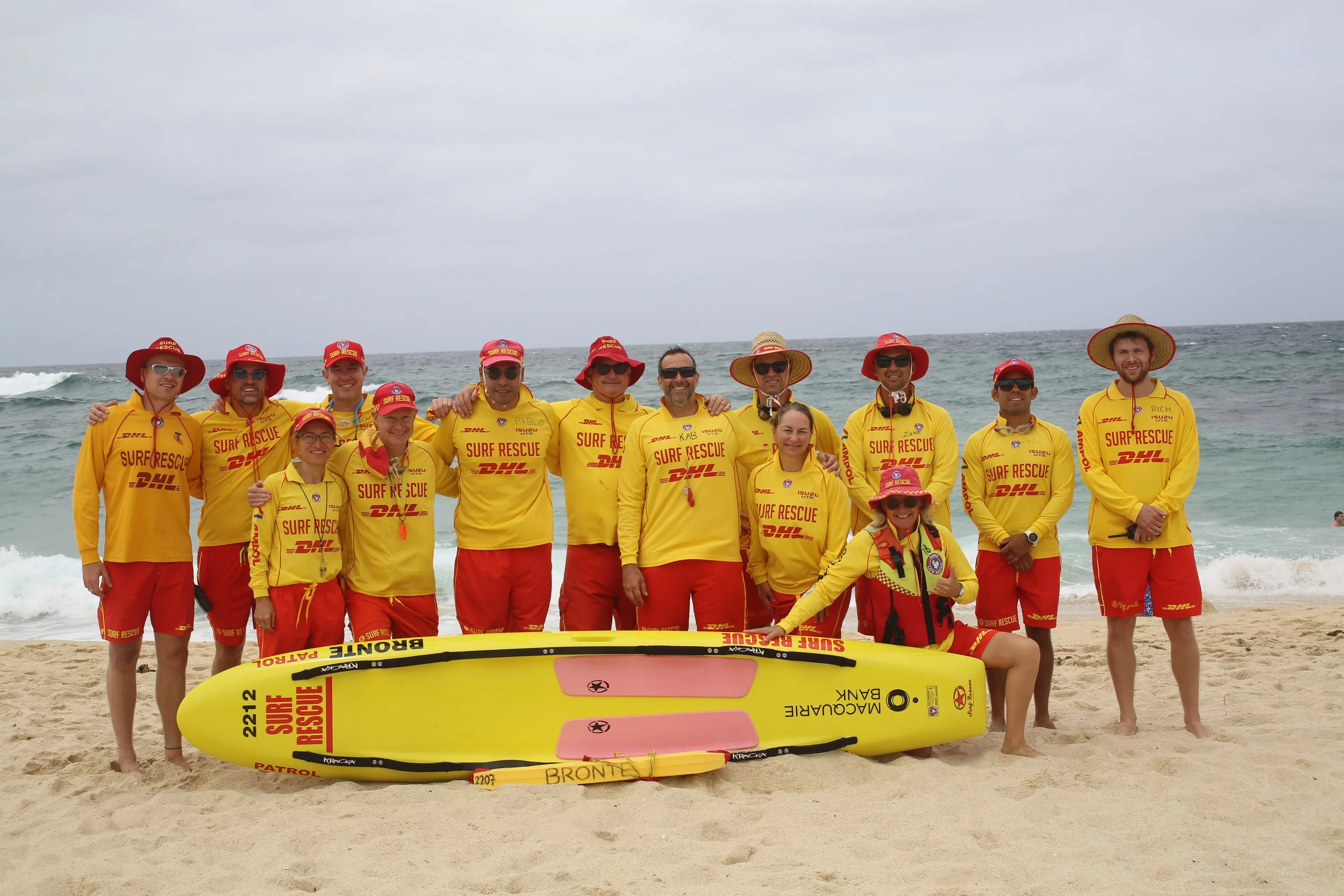 A group of people dressed in yellow surf rescue uniforms standing on a beach with surf rescue equipment, smiling at the camera. The background shows the ocean and cloudy sky.