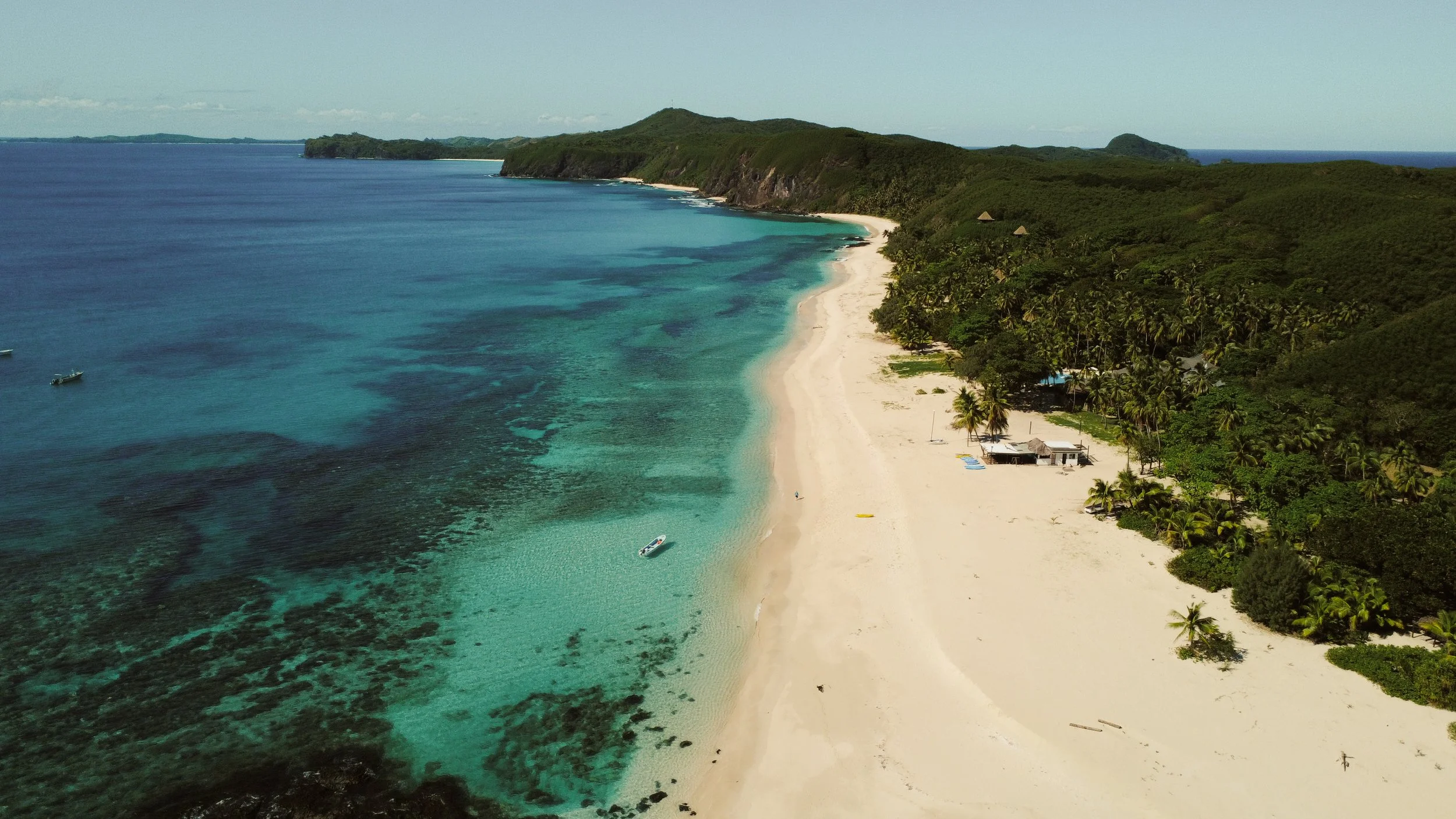 Aerial view of a tropical beach with white sand, turquoise water, lush green hills, and a few small boats near the shore.
