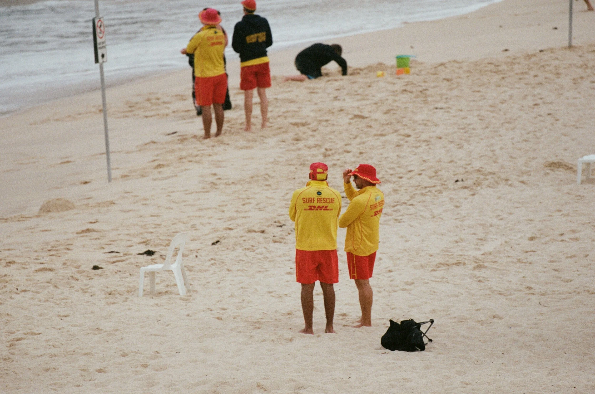 Two people in yellow firefighter uniforms and red hats are standing on a sandy beach, talking, with two other individuals nearby. One of those individuals is placing something in the sand while another observes. Toward the water, two people are stand
