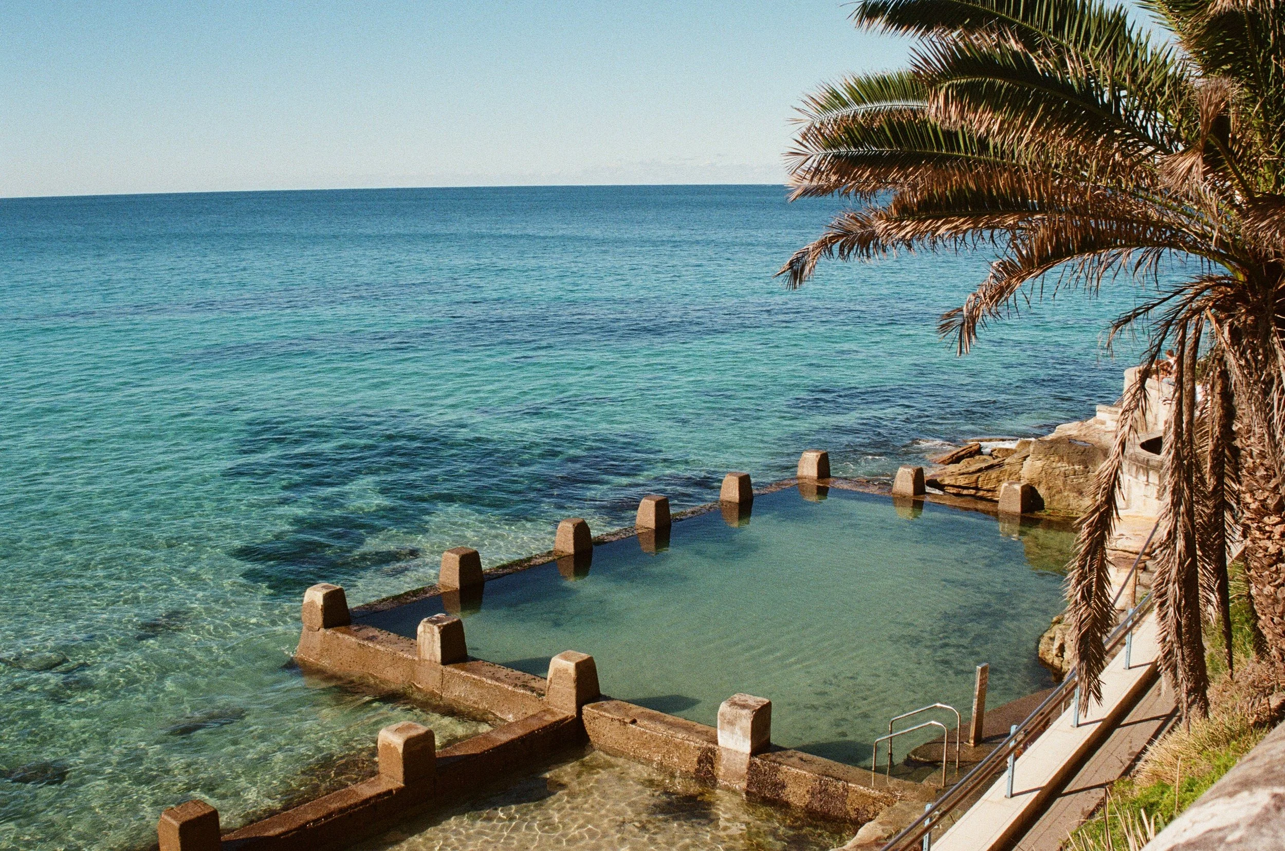 Ocean with greenish-blue water, a palm tree on the right, rocks and a small pool with steps leading into the water.