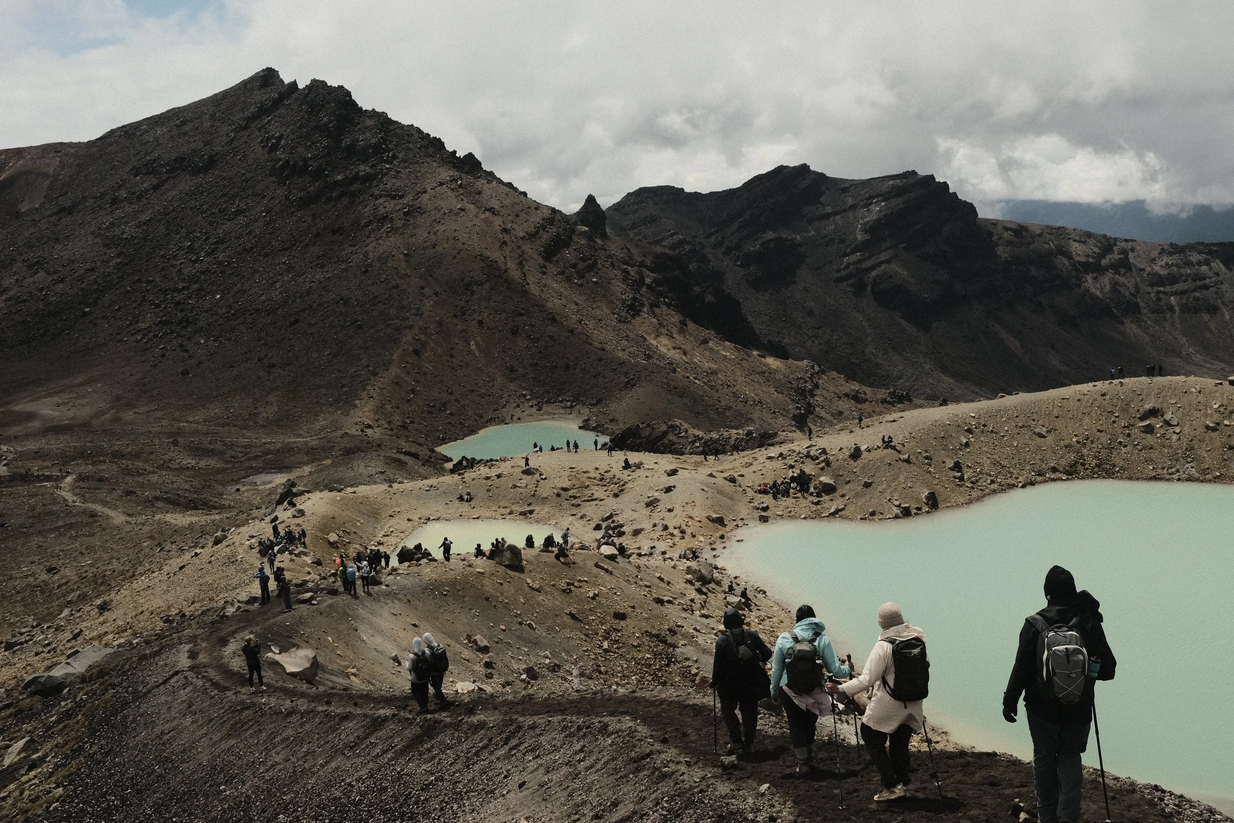 Group of hikers walking along a rocky path near turquoise crater lakes in a volcanic landscape under cloudy sky.