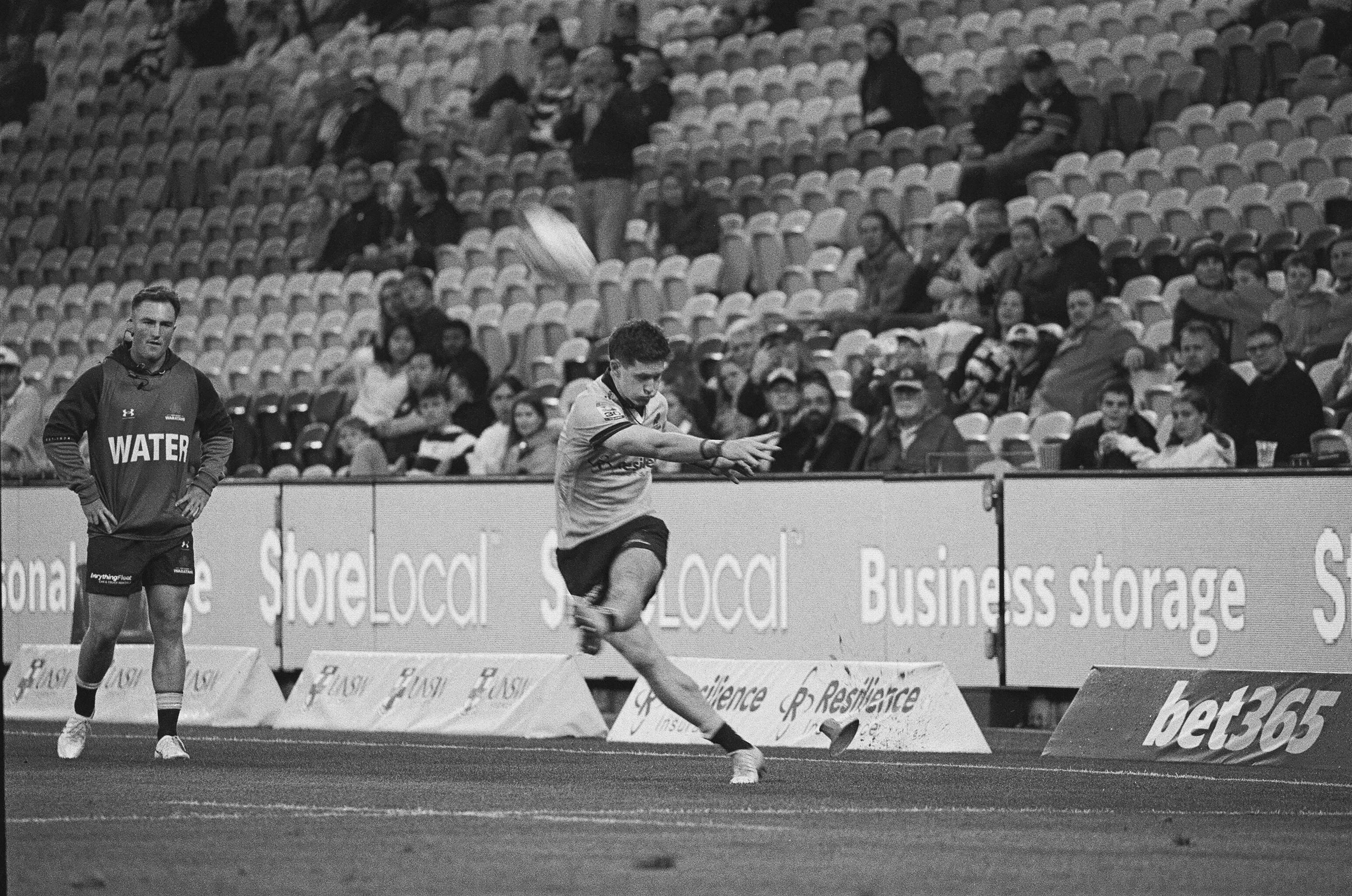 A black and white photo of a soccer match with a player kicking the ball and another standing nearby, while spectators watch from the stands.
