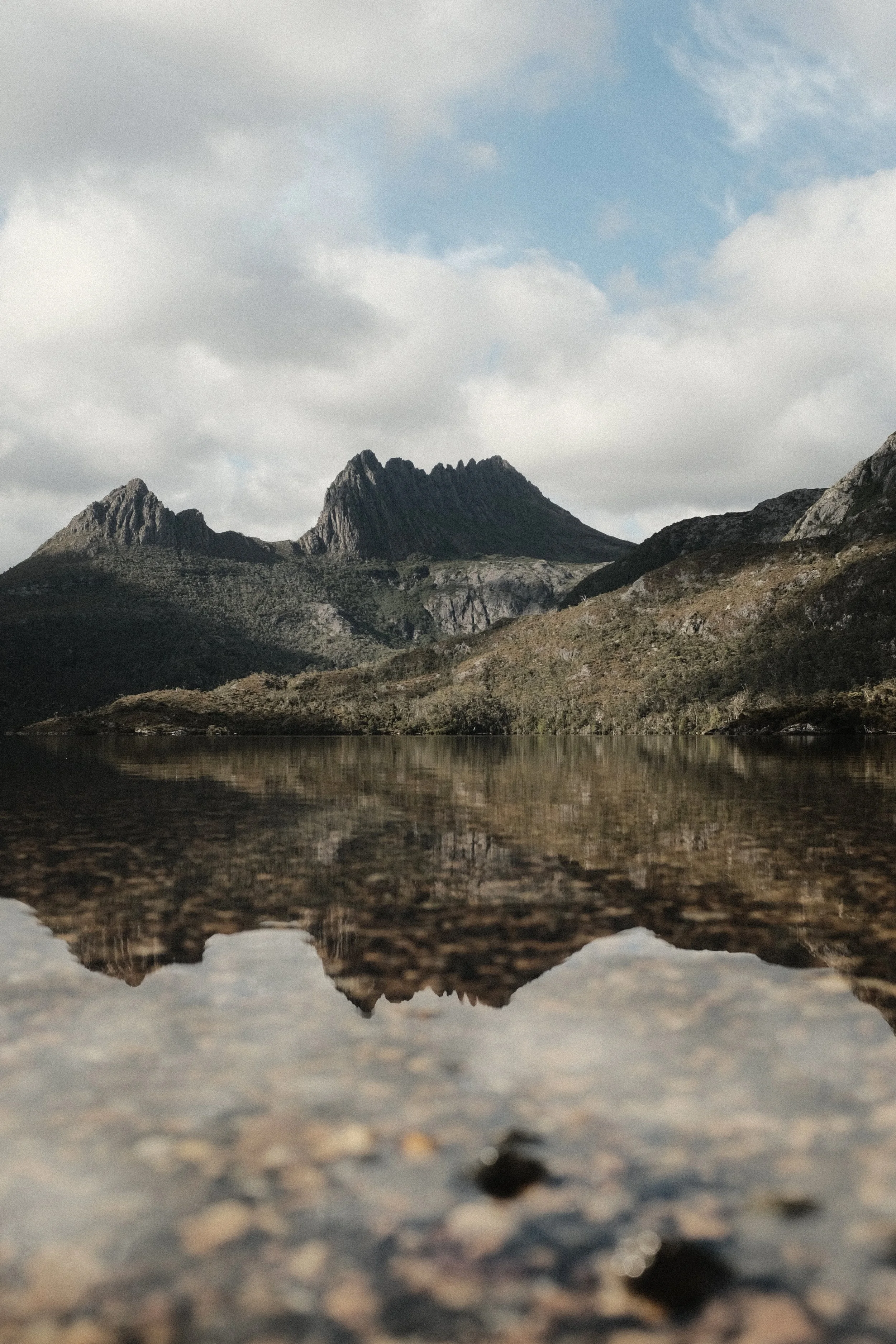 Mountain range reflected in a calm body of water with rocky and grassy terrain in the foreground, cloudy sky overhead.