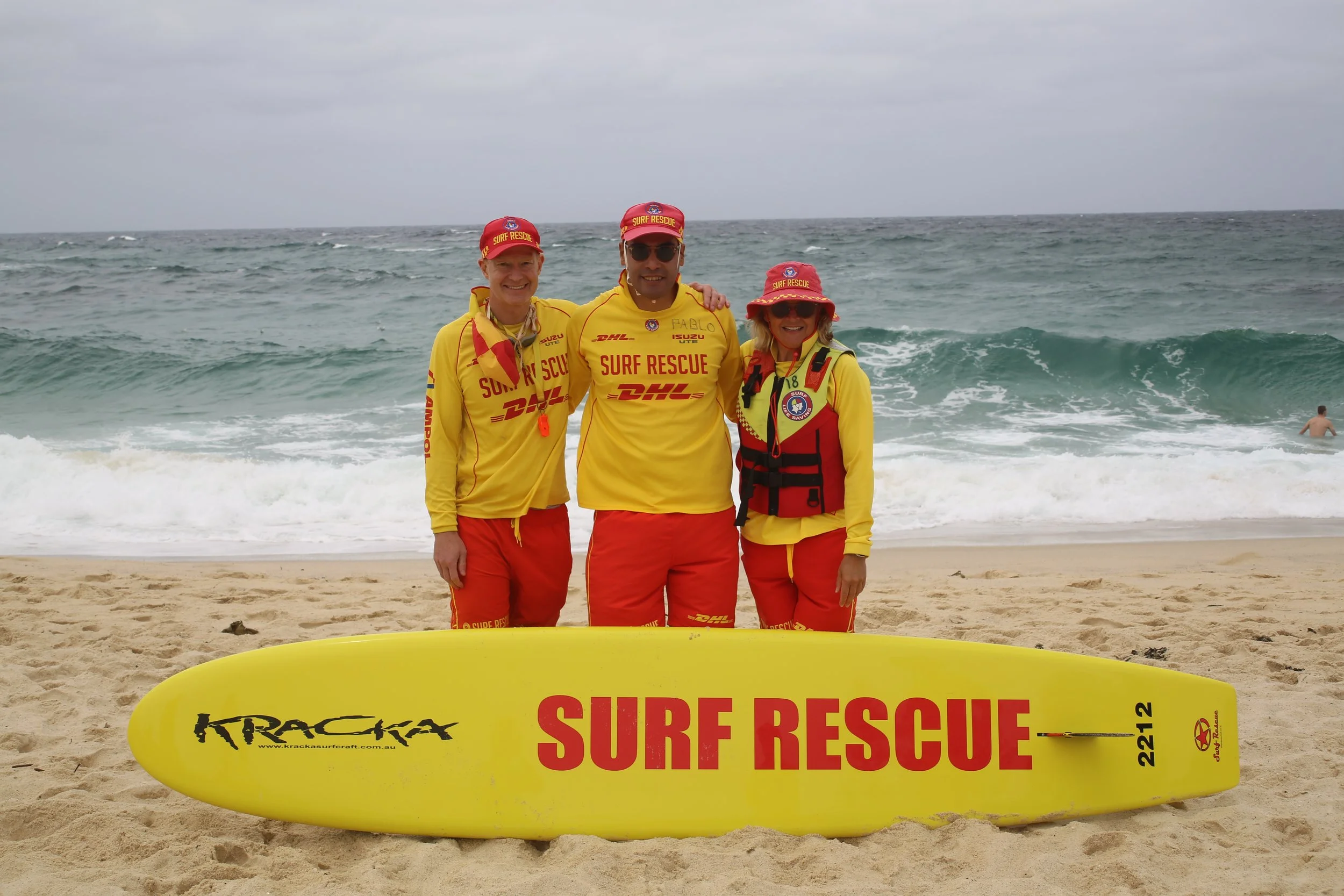 Three surf rescue team members dressed in yellow and red uniforms standing on the beach with a yellow rescue board, ocean waves in the background, and an overcast sky.