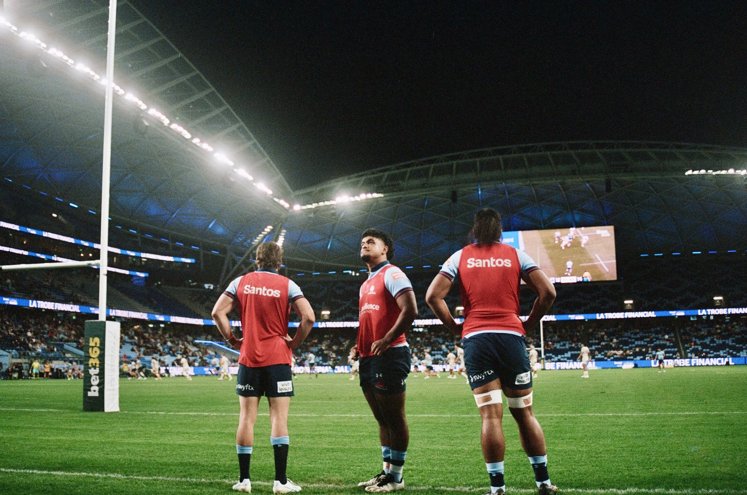 Three rugby players in red and blue uniforms are standing on a green field inside a stadium at night, watching the game. The stadium is filled with spectators and large screens are visible in the background.