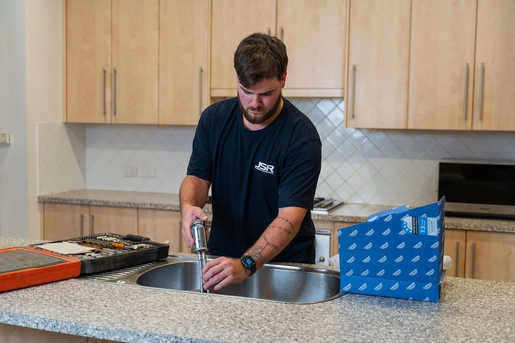A man working on a plumbing repair at a kitchen sink, with tools and a blue delivery box nearby.