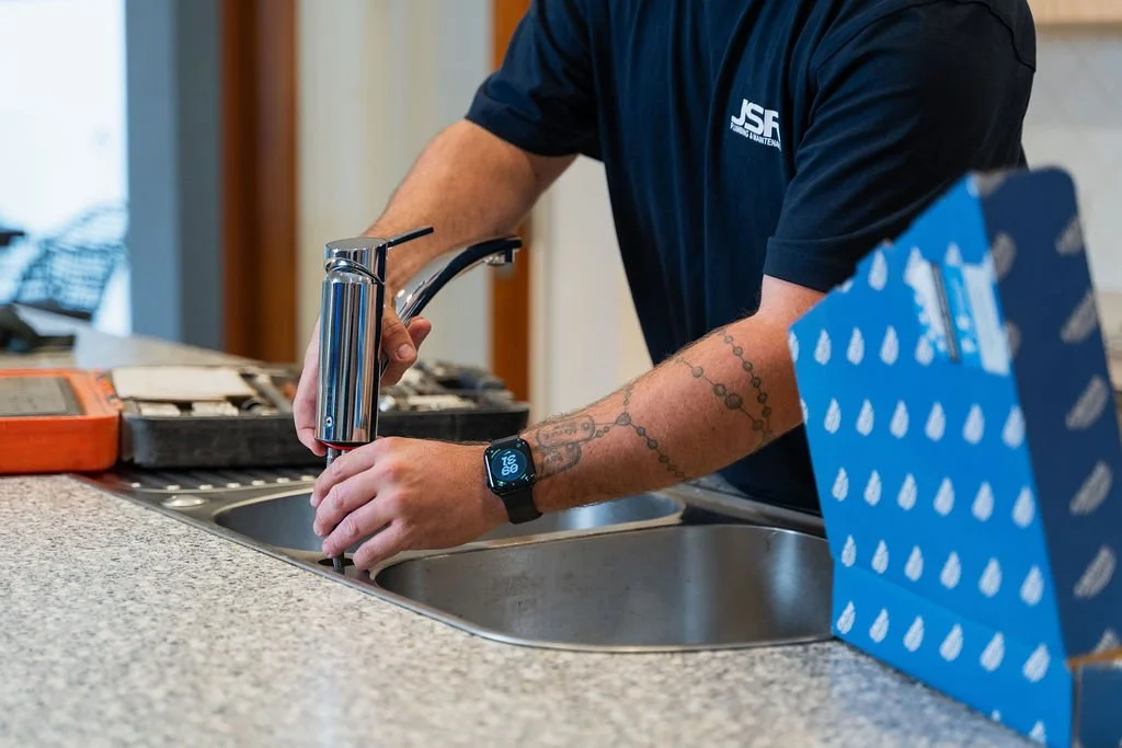 Person filling a bottle at a kitchen sink with a modern faucet, wearing a blue shirt and smartwatch, with a blue box on the counter.