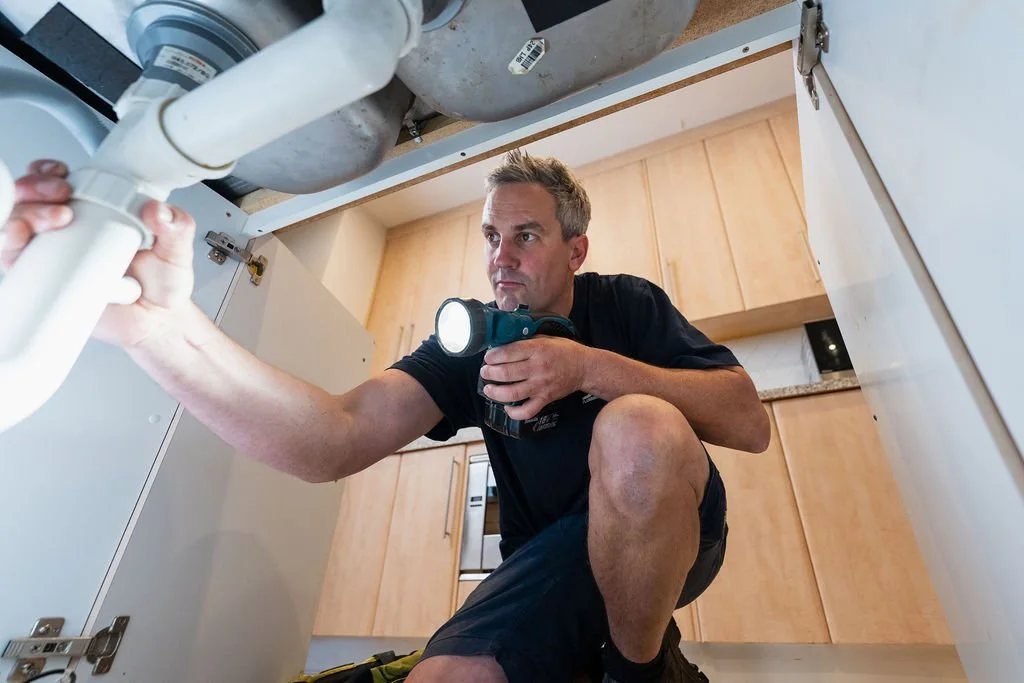 A man in a black shirt working on plumbing under a kitchen sink with a flashlight.