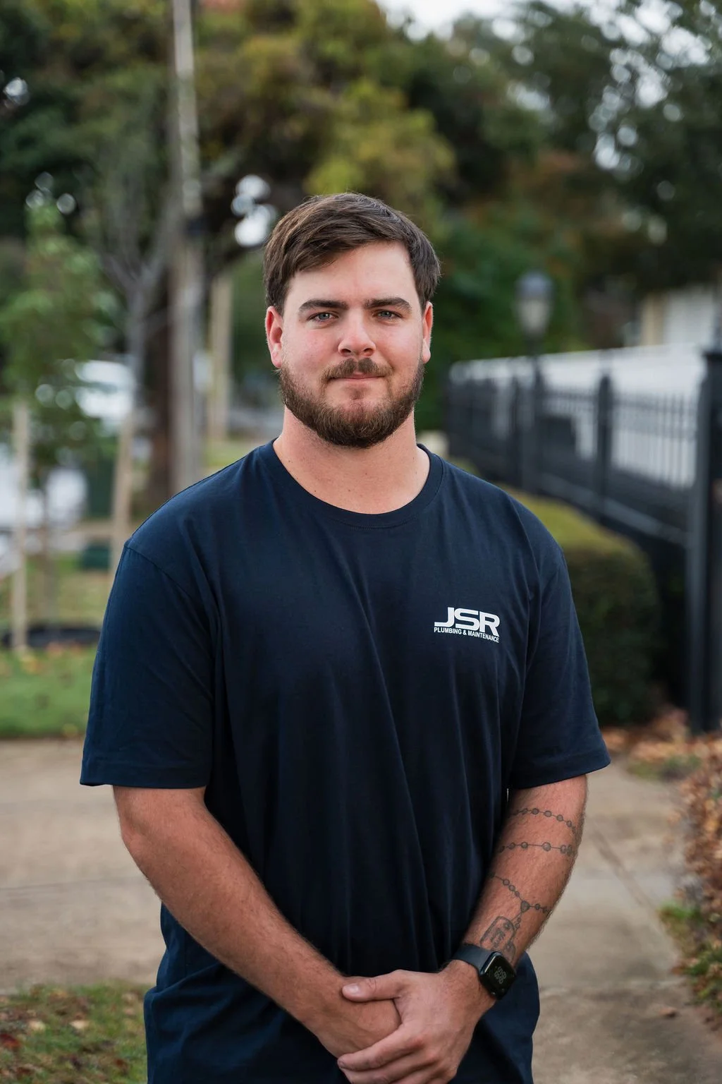 A young man with dark hair, a beard, and light skin, wearing a navy blue T-shirt with "JSR Plumbing & Maintenance" logo, standing outdoors on a sidewalk with trees and a black fence in the background.