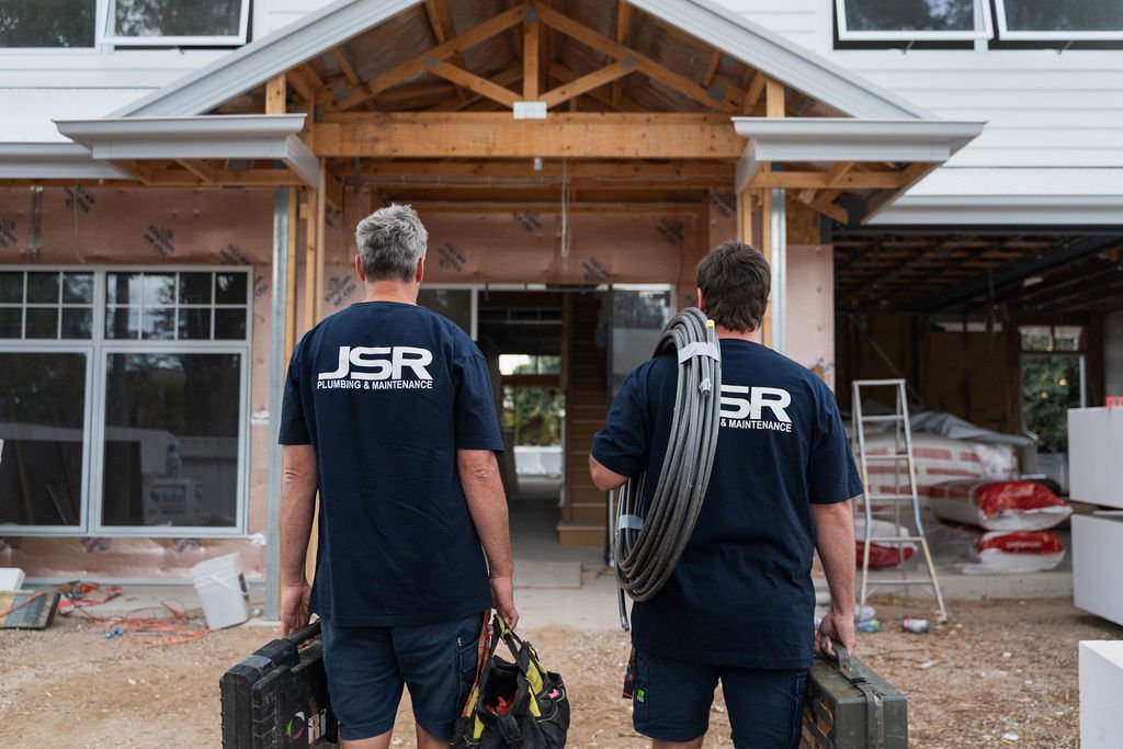 Two workers with tools walking towards a house under construction, wearing matching shirts that say 'JSR Plumbing & Maintenance'