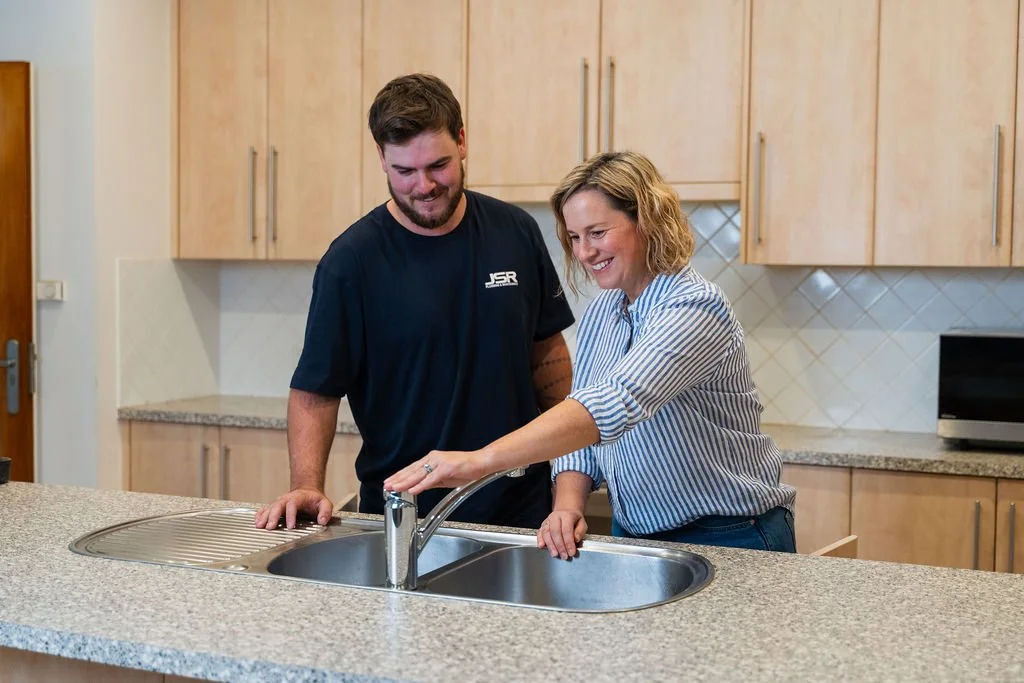 A woman and a man smiling in a kitchen, standing at a sink, with the woman turning on the tap.