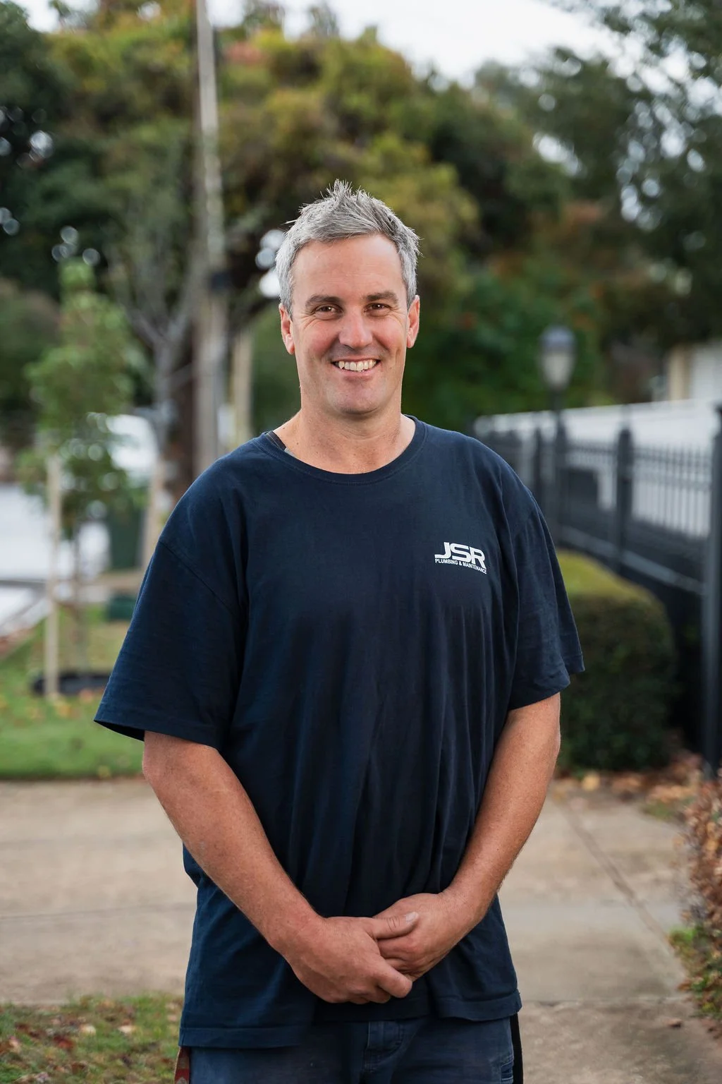 A smiling man with gray hair standing outdoors on a sidewalk with trees and a fence in the background. He is wearing a dark blue t-shirt with white text on it.