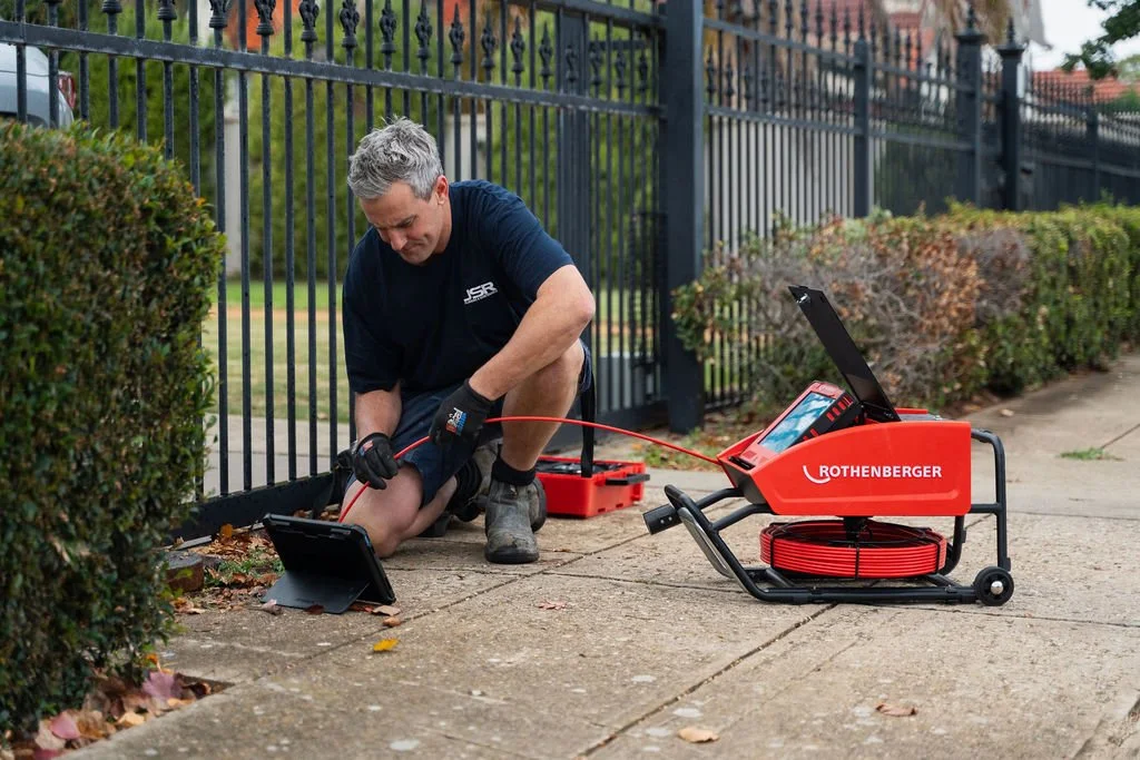 A man is crouching on the sidewalk near a black metal fence, using a diagnostic device connected to a red Rothenberger machine by a red cable, with a tablet propped on the ground nearby.