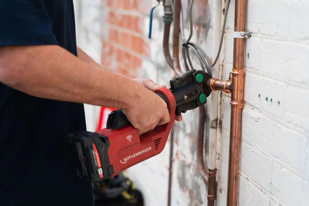 A person using a red Rothenberger pipe threading machine on copper pipes against a white brick wall.