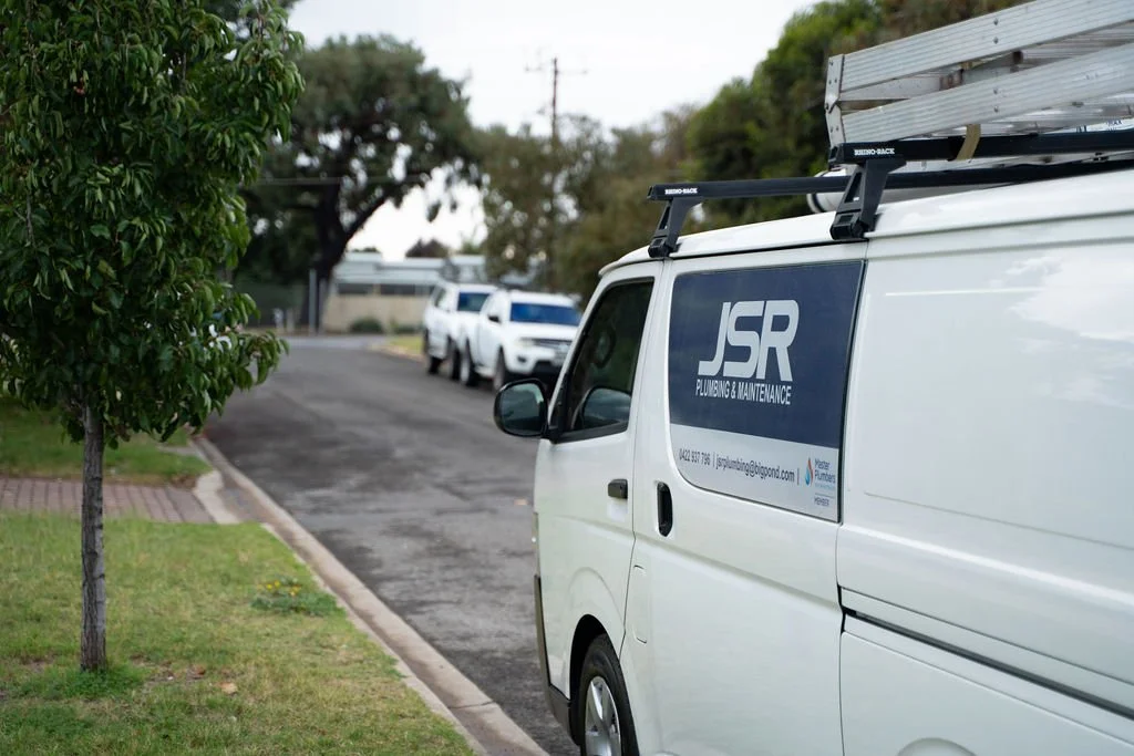 White utility van with JSR Plumbing & Maintenance logo parked on a suburban street, trees, and other vehicles in the background.